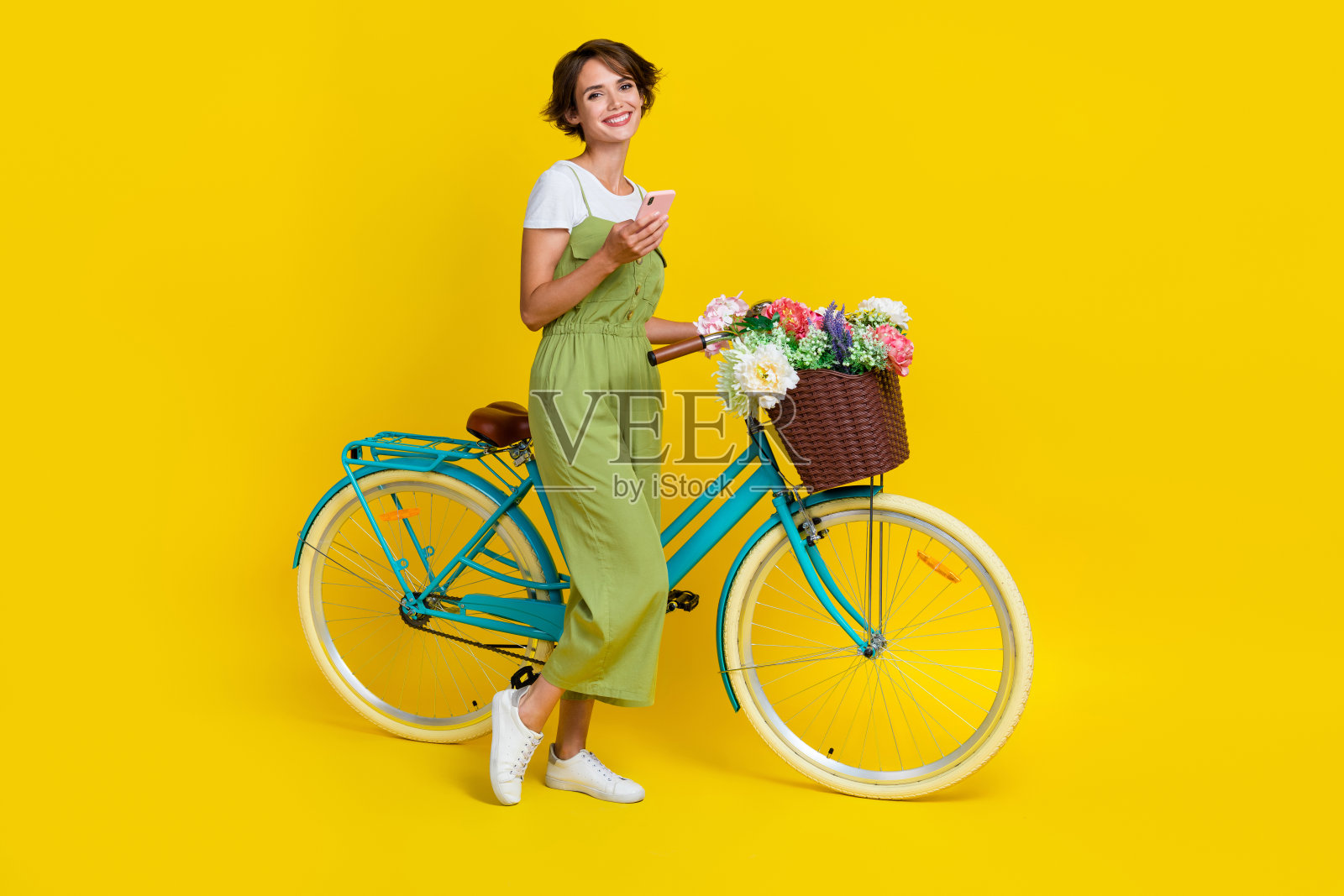 全身照：年轻漂亮的女性手持设备，骑自行车，走路时提着花篮，穿着时尚的卡其色服装，背景为黄色。照片摄影图片
