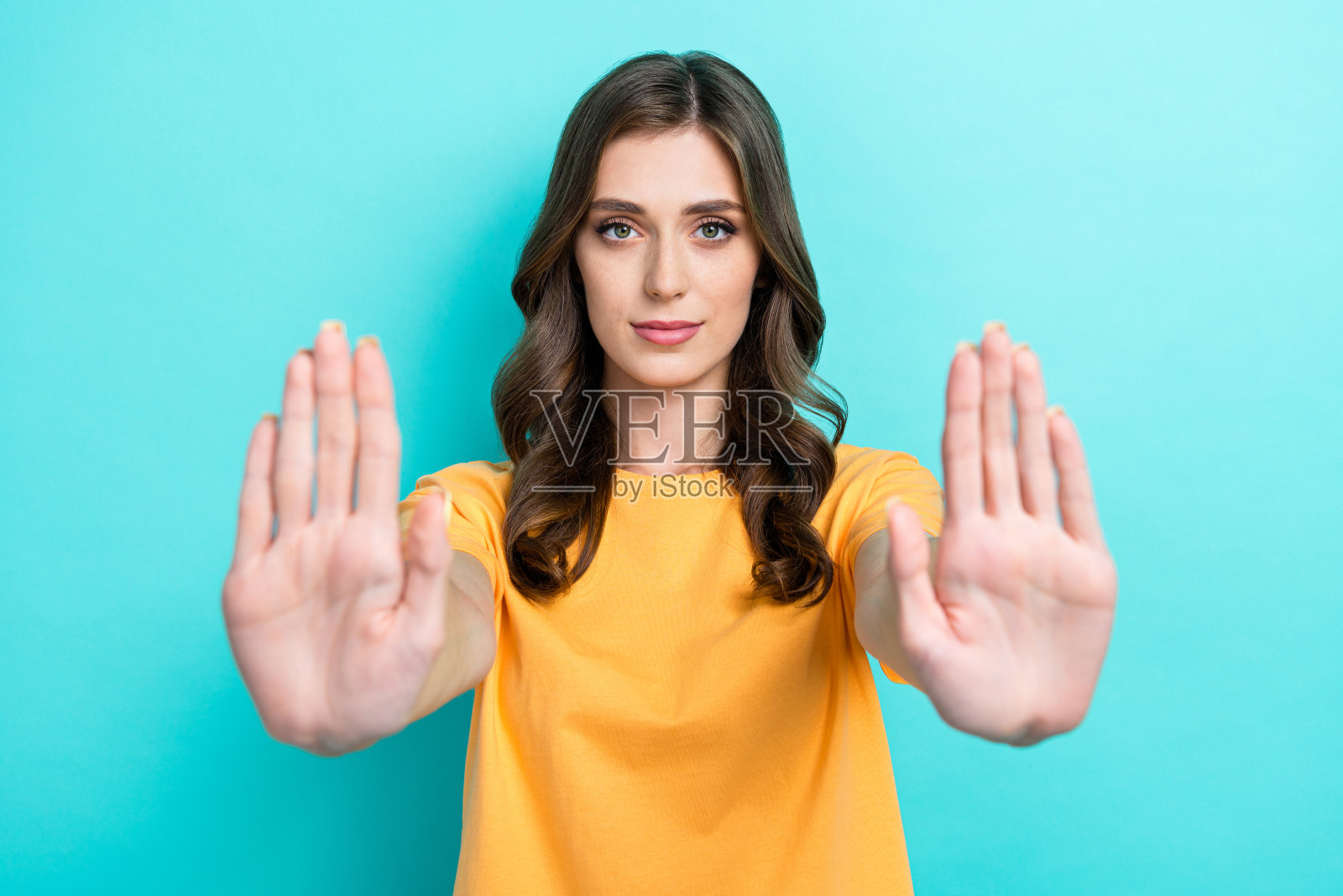 自信的漂亮女士穿着黄色T恤，双手掌心向外，表示停止，背景为青绿色。照片摄影图片