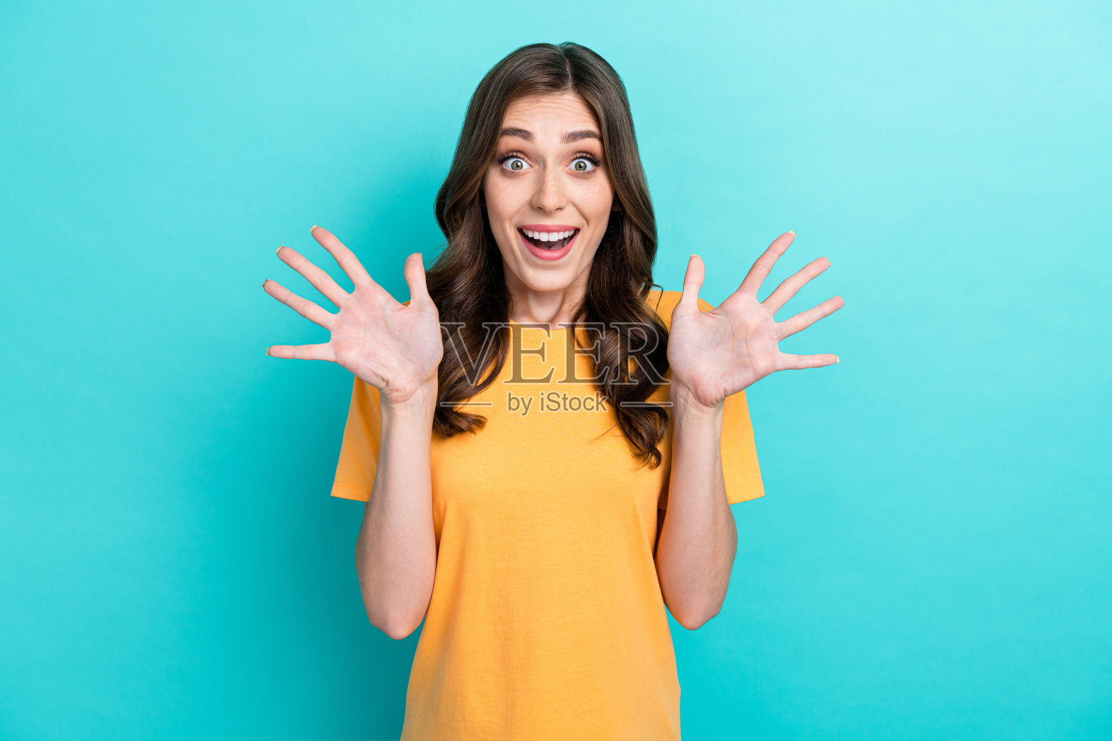 年轻可爱漂亮的女孩穿着黄色T恤，惊讶地用十根手指指着，兴奋而意外，背景为青色。照片摄影图片
