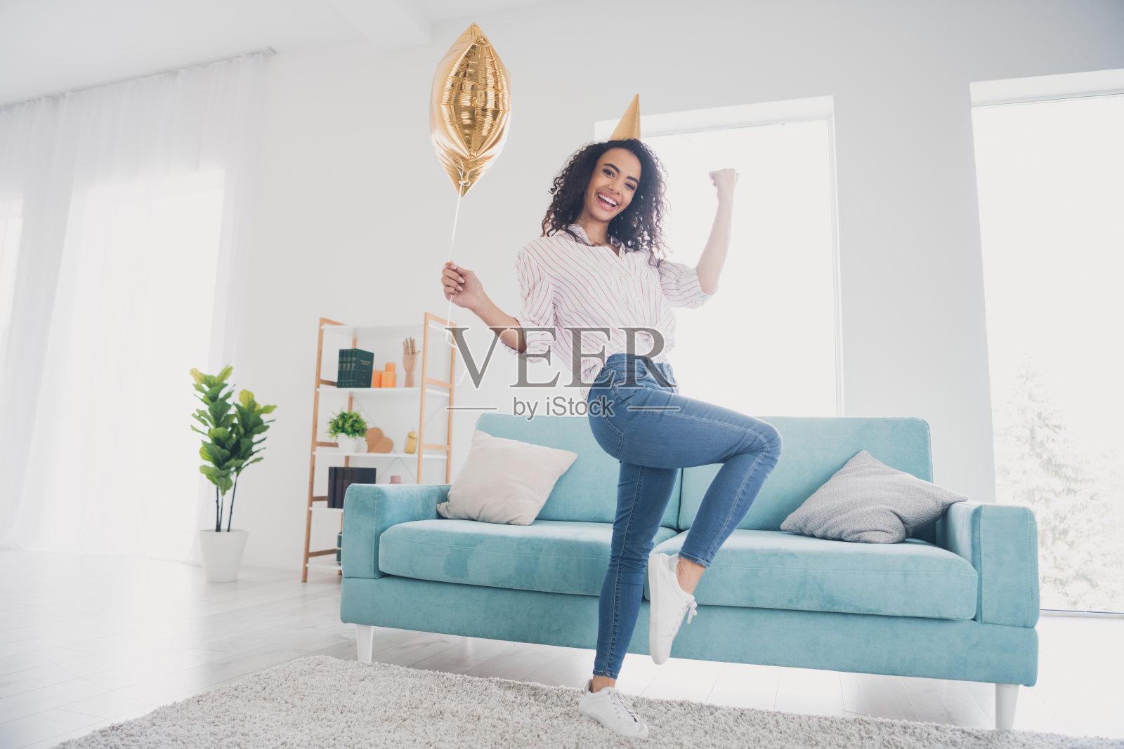 全身肖像的漂亮年轻女孩举起拳头，戴着尖顶帽，手持星形气球，庆祝生日派对，室内平面图。照片摄影图片