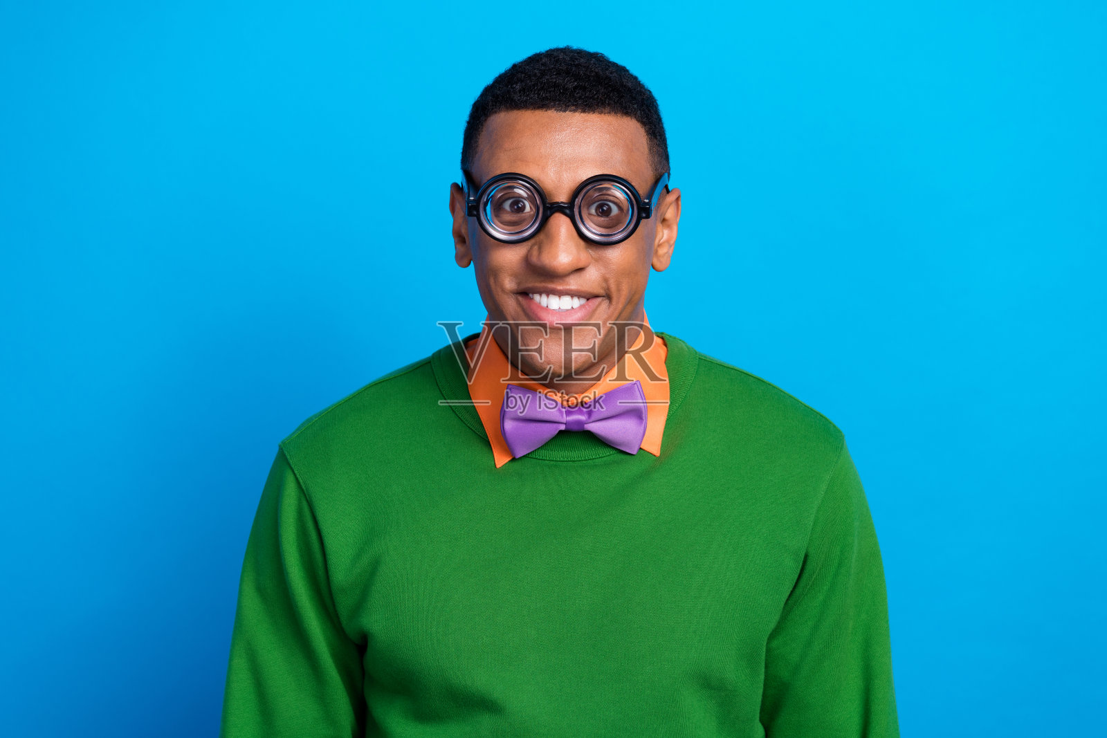 有趣兴奋的男士肖像照，露出灿烂的笑容，佩戴着时尚的紫色蝴蝶结和眼镜，显得童趣十足，背景为蓝色。照片摄影图片