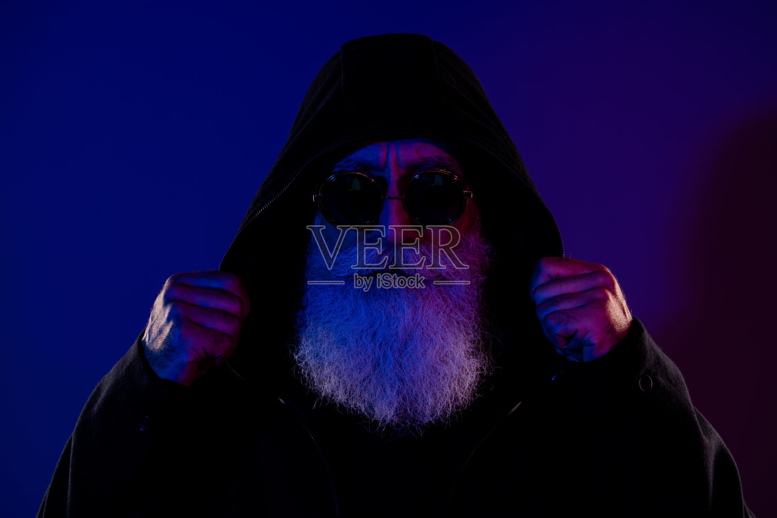 神秘酷炫的退休男士，穿着黑色套头衫，戴着眼镜，双臂交叉，头巾遮住自己，背景为深蓝色。照片摄影图片