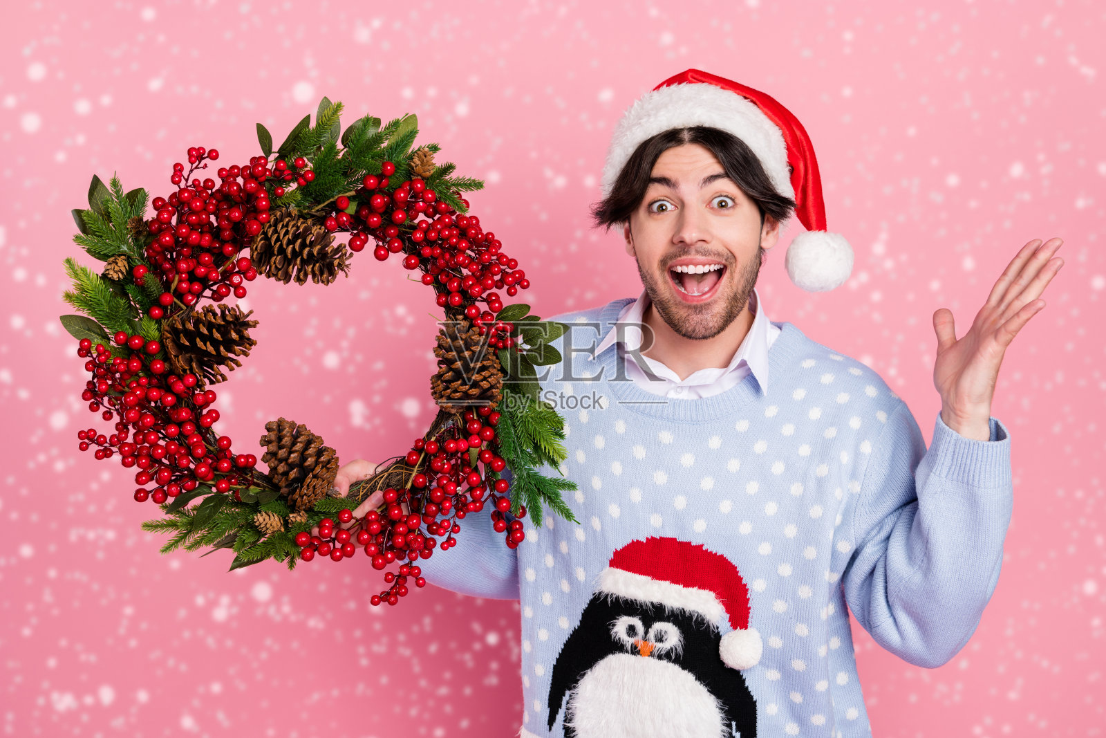 兴奋的幽默绅士穿着新年毛衣，微笑着举起手臂，手中拿着装饰品，背景为粉色。照片摄影图片
