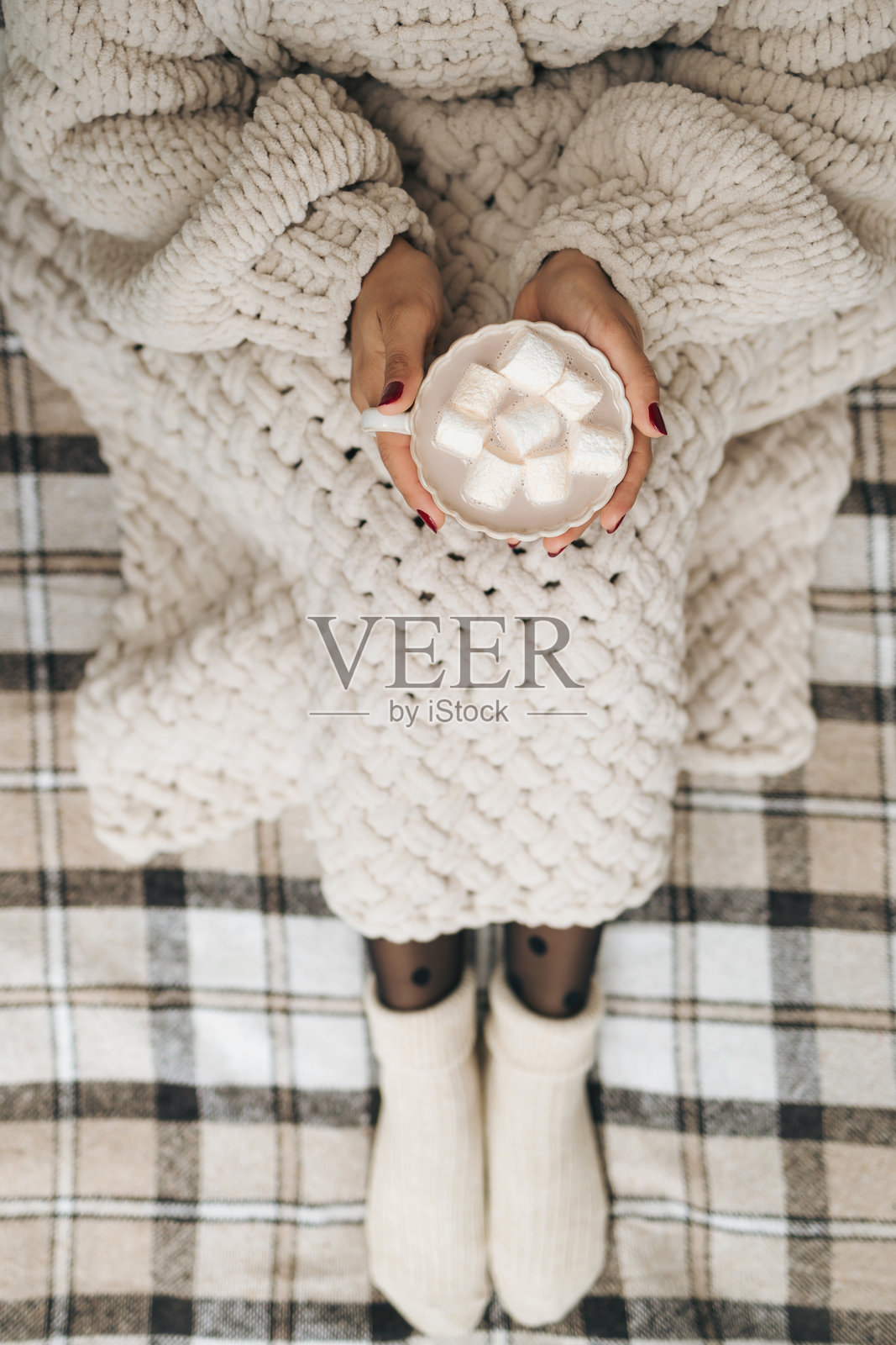 穿着针织毛衣的女人坐在舒适的格子毯子上，手里拿着一杯热巧克力或可可饮料，上面放着棉花糖。温馨舒适的生活理念。照片摄影图片