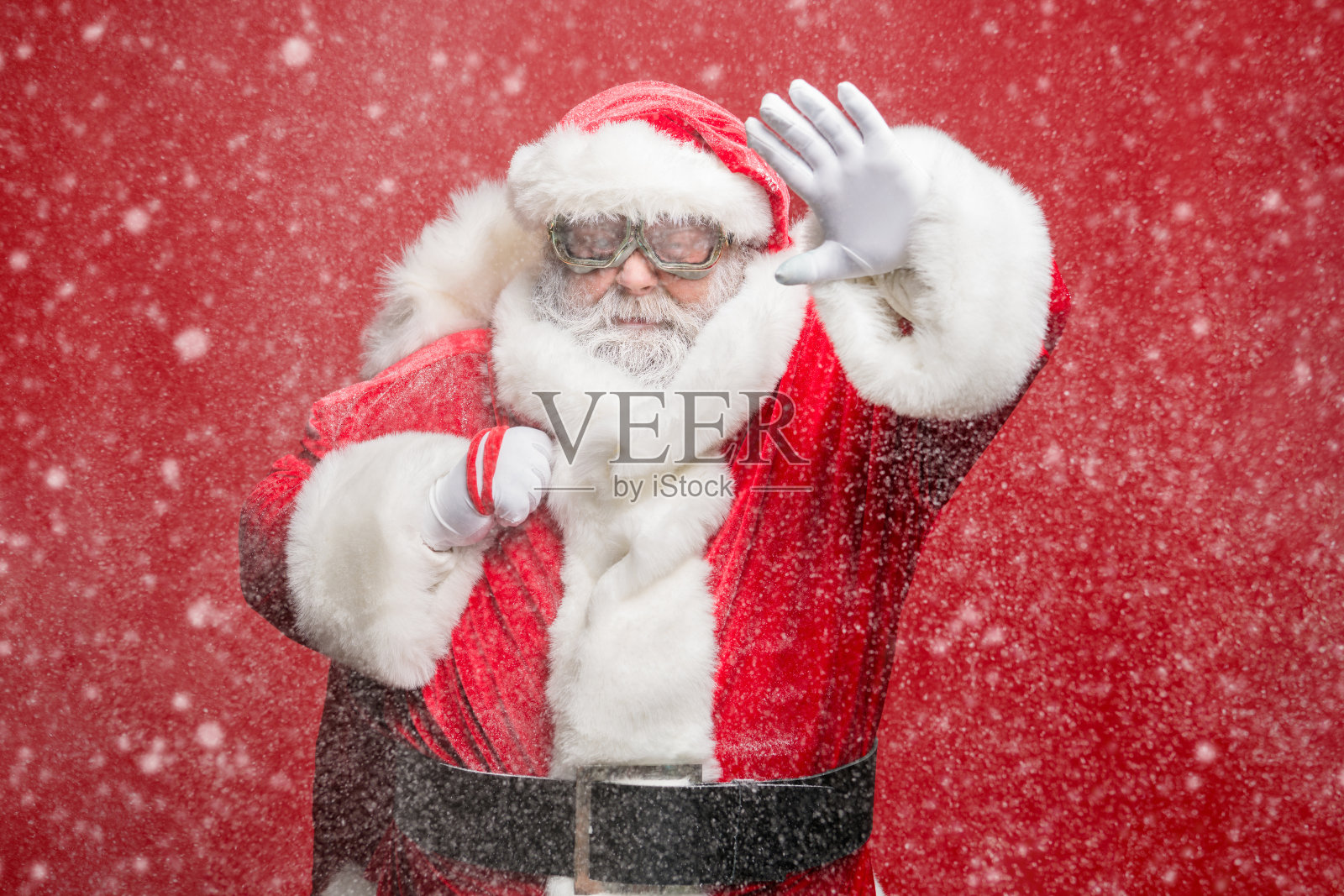 强烈的自然灾害，注意！一位不快乐的退休老人穿着圣诞老人服装，面临能见度问题，看到雾霭，背着袋子，戴着帽子和护目镜，孤立在红色背景上。照片摄影图片