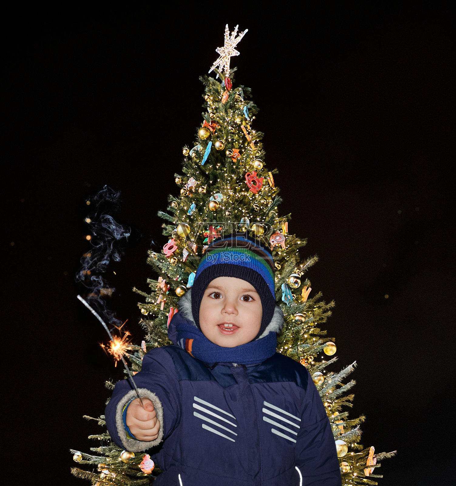 一个快乐的孩子在美丽装饰的圣诞树前手持烟花棒。照片摄影图片