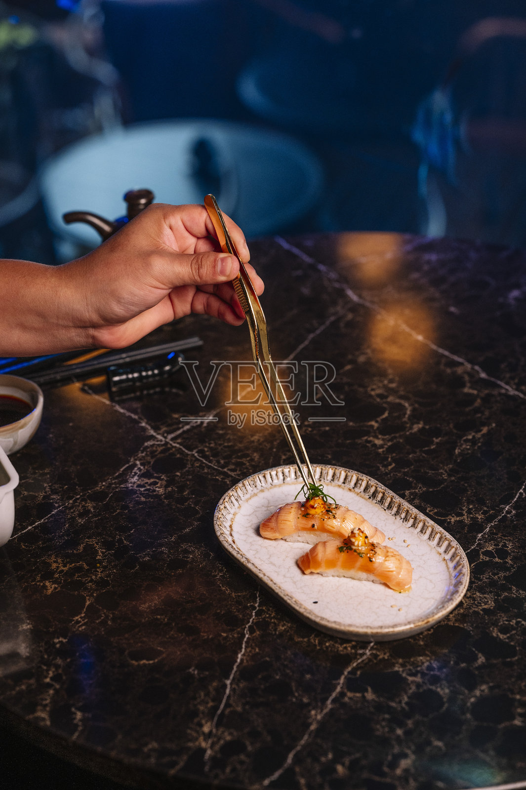 特写镜头显示一只手用金色夹子小心翼翼地夹起一块寿司。照片摄影图片