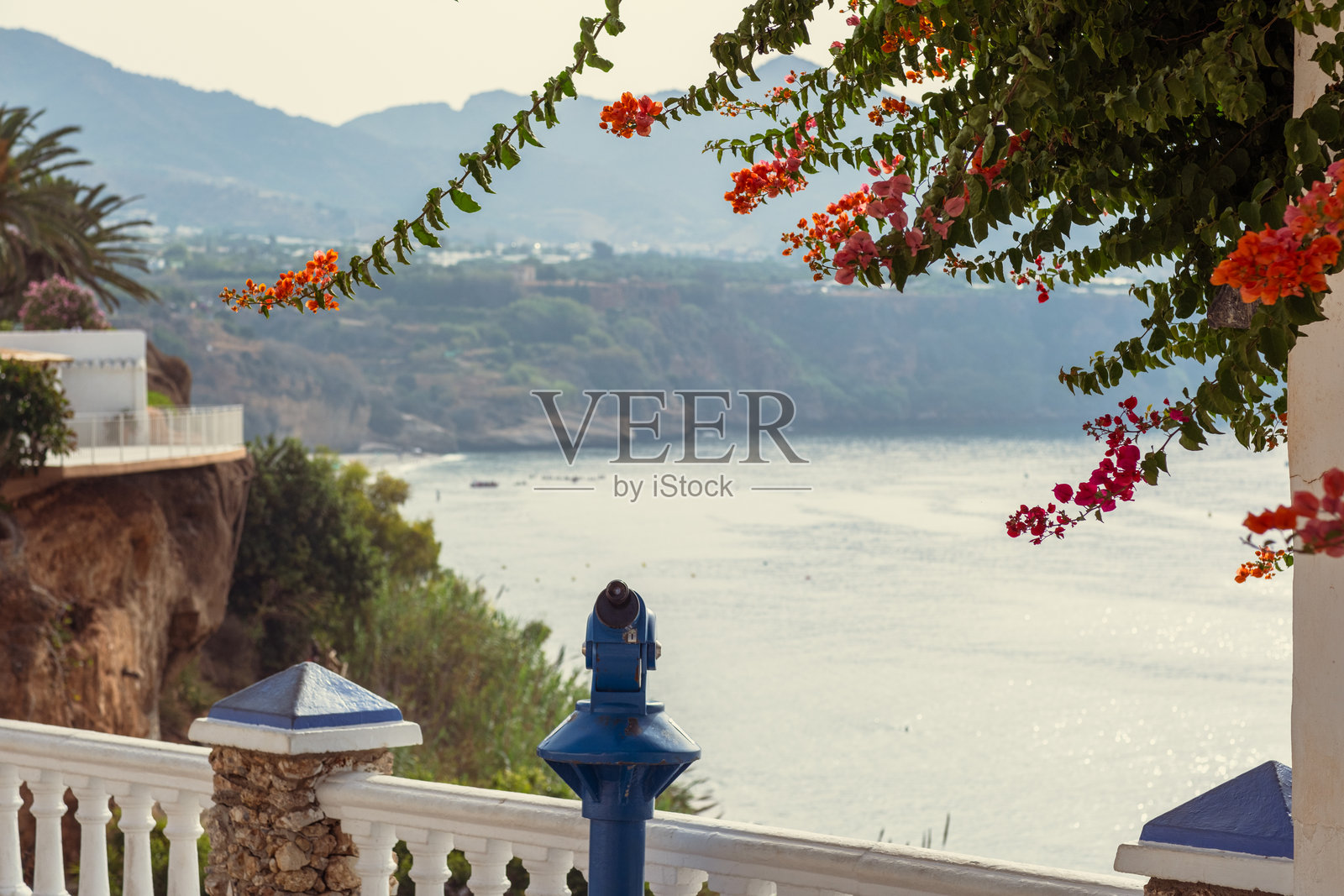 西班牙的海滨度假胜地。前景是红色花朵和一个带望远镜的观景平台，背景是模糊的海景。照片摄影图片