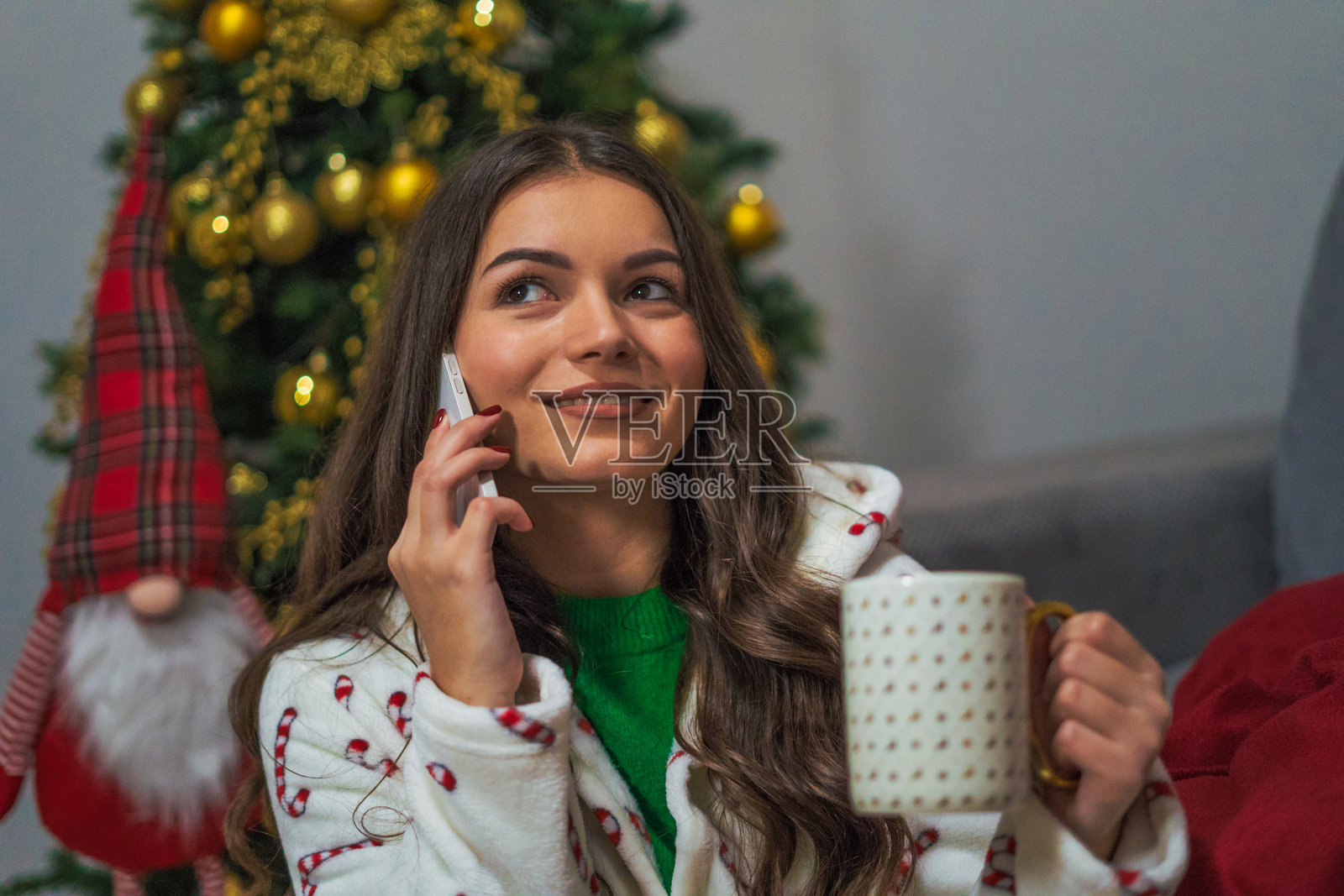 一位年轻、快乐的女性在她装饰一新的家中，穿着圣诞主题的浴袍，站在树前打电话。照片摄影图片