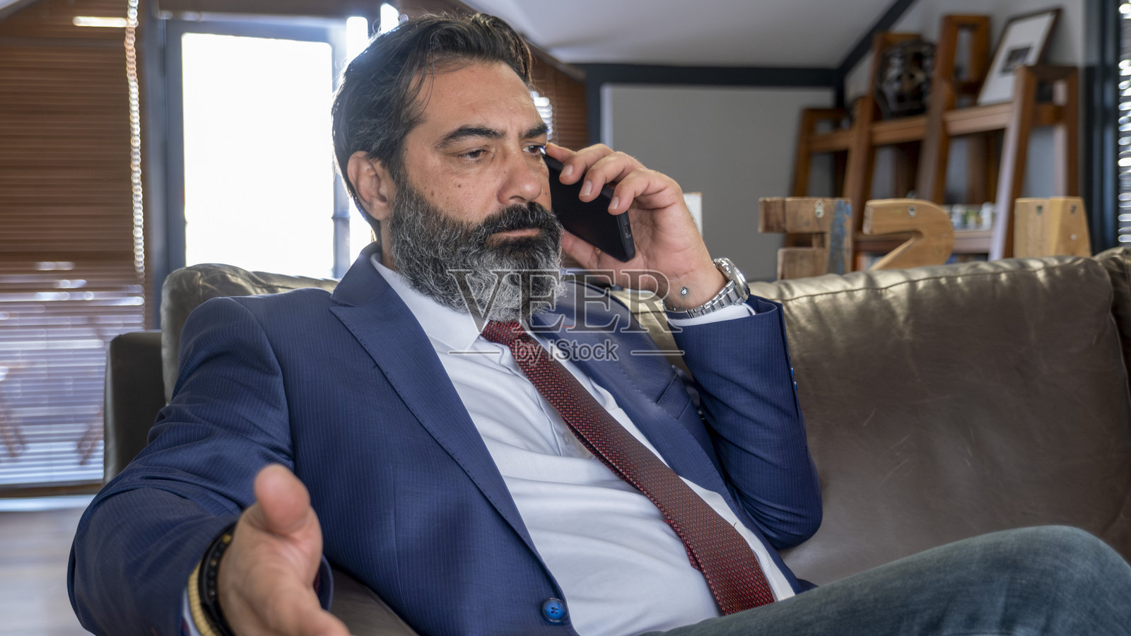 一位中年黑发商人在办公室的皮沙发上坐着，正在通过电话进行紧张的商务会议。照片摄影图片