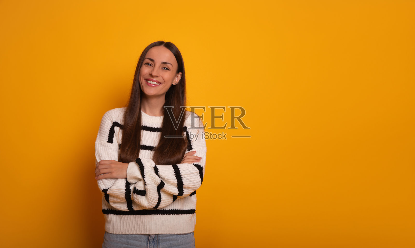 一位微笑的女性穿着休闲的白色毛衣，自信地站在明亮的橙色背景前，双臂交叉。她看起来放松且亲切。生活方式或宣传内容。照片摄影图片