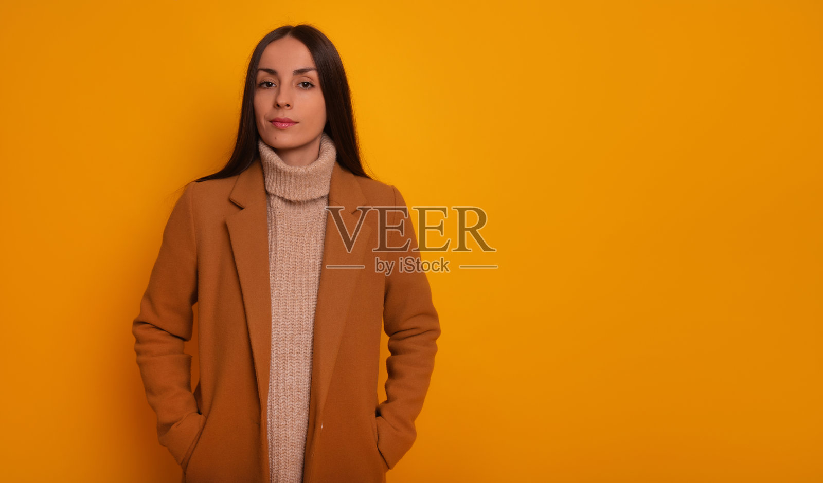 一位穿着驼色大衣和米色高领毛衣的时尚女性自信地凝视着镜头，背景是明亮的黄色。她在这张肖像照中散发出冷静、优雅和时尚的气息。照片摄影图片