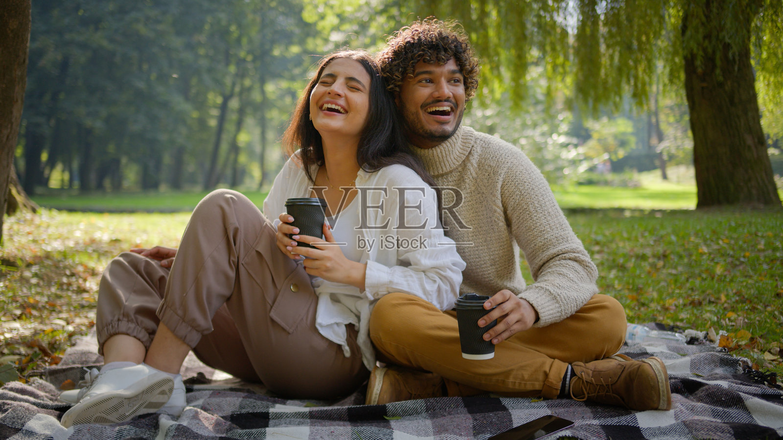 快乐的多民族已婚情侣，男士和女士在户外公园的草地上坐在毯子上，手里拿着一次性咖啡杯，享受着浪漫的约会时光。照片摄影图片