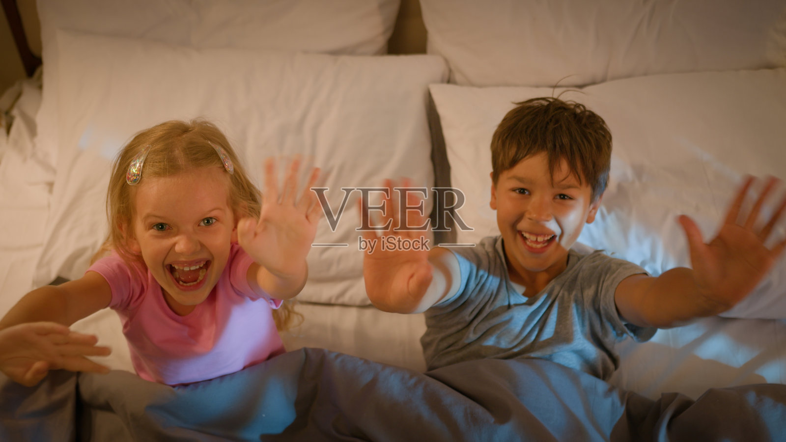 两个快乐、微笑、有趣、友好的白人孩子，兄弟和姐妹，躺在床上，枕头上，惊喜地挥手打招呼，晚上在室内卧室的毯子下。照片摄影图片