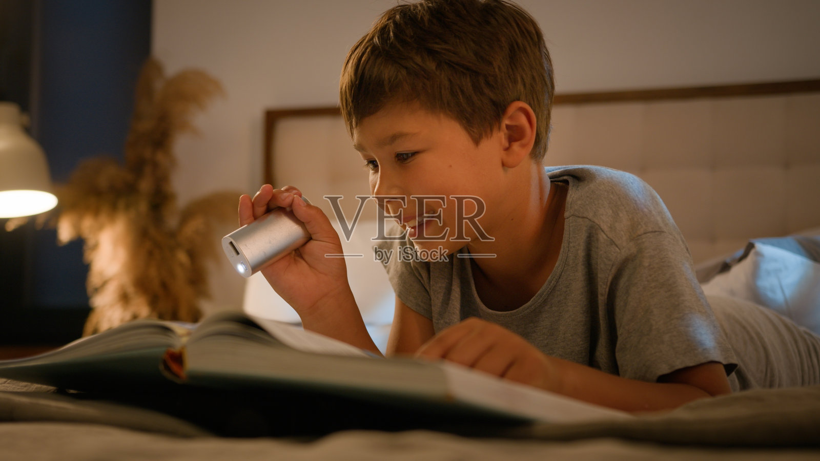 惊讶、开心、搞笑的白人男孩在卧室的床上用手电筒躺着阅读一本有趣的书，享受着夜晚的睡前童话故事。照片摄影图片