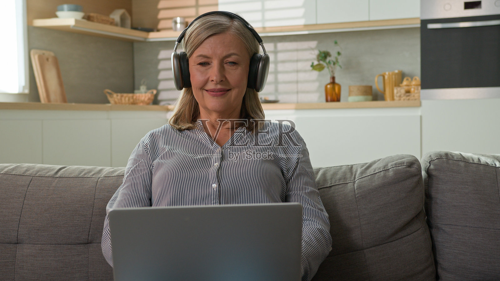 微笑的成熟白人女性祖母，作为自由职业者在家中使用笔记本电脑打字，戴着耳机听音乐，交流社交网络，现代科技与老年人的概念。照片摄影图片