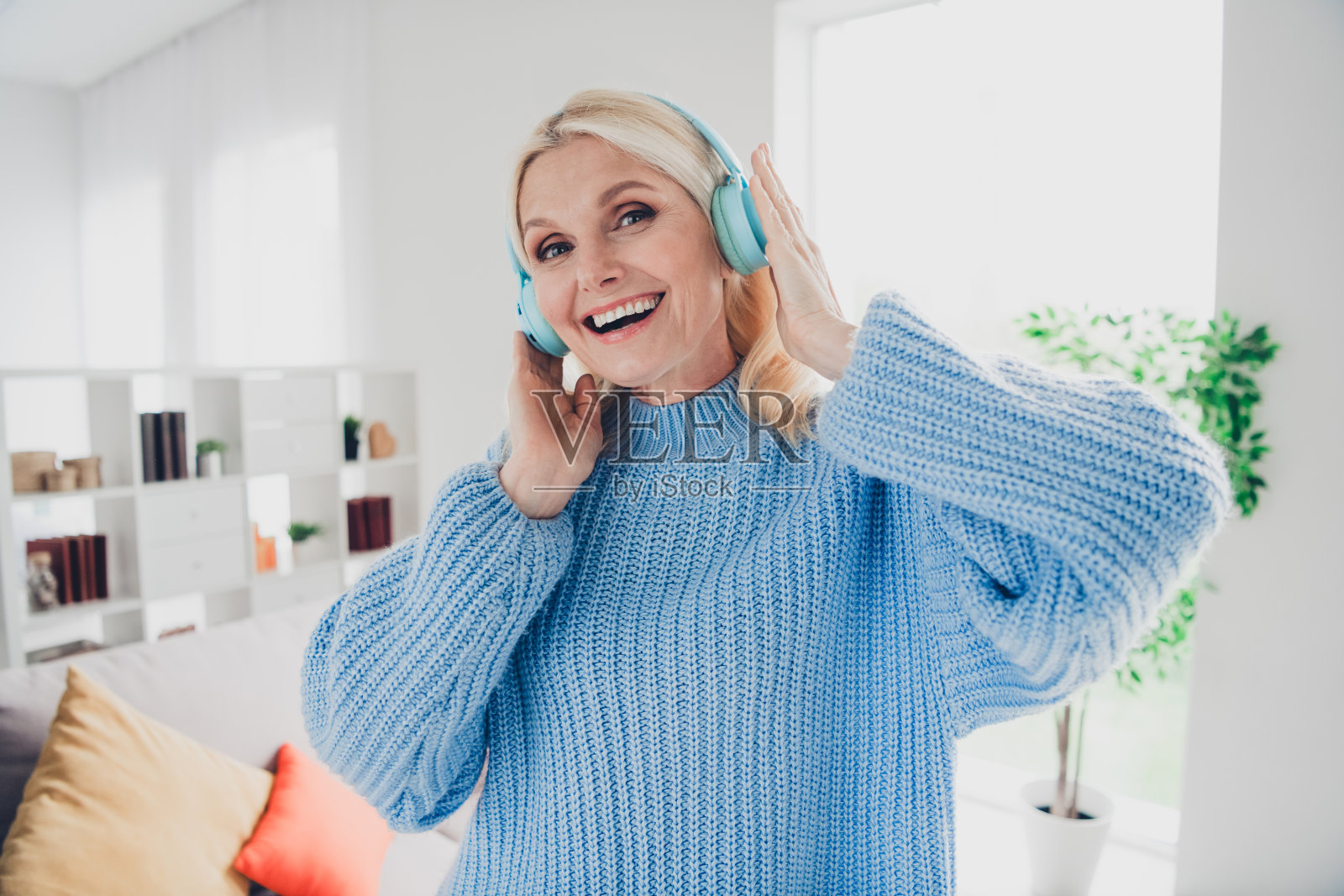 微笑的老年女士，戴着耳机，穿着毛衣，享受周末的休闲时光，坐在现代公寓室内。照片摄影图片