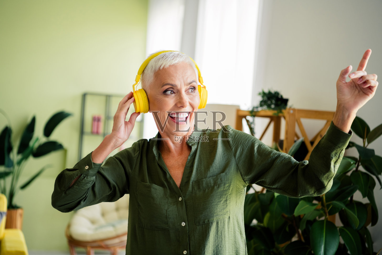 穿着绿色衬衫的漂亮快乐女士在室内家中戴着耳机听歌跳舞照片摄影图片