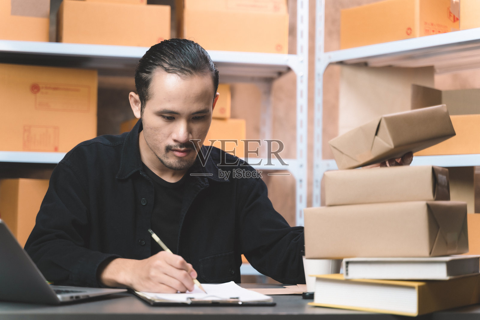 一名工作男士在笔记本电脑上查看在线订单并做笔记。他在家中的办公室和小商店里从事新的在线购物业务。照片摄影图片