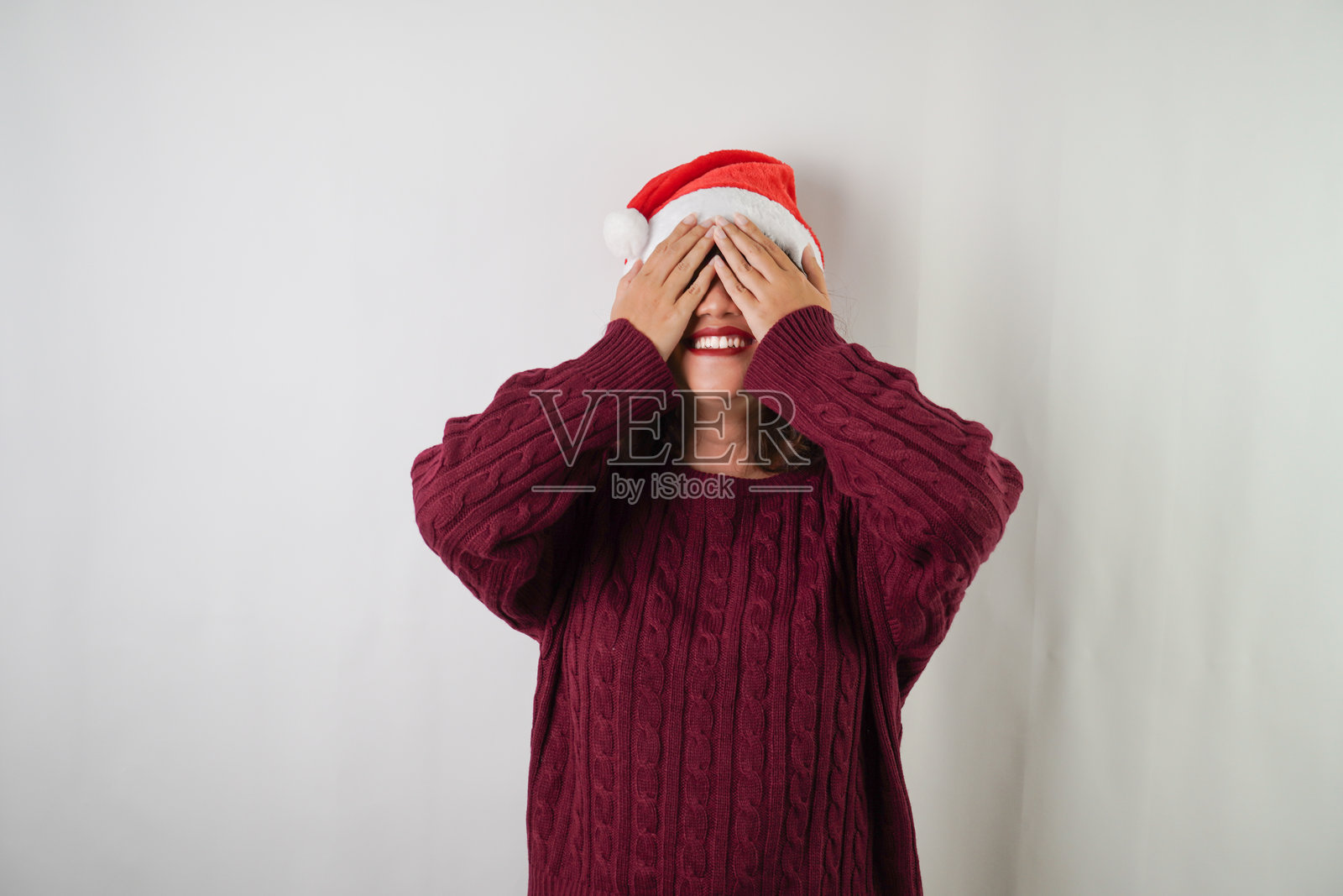 年轻的亚洲女性戴着圣诞老人帽，穿着红色长袖毛衣，用手掌遮住脸，背景为白色。适合圣诞节假期和新年派对的概念。照片摄影图片
