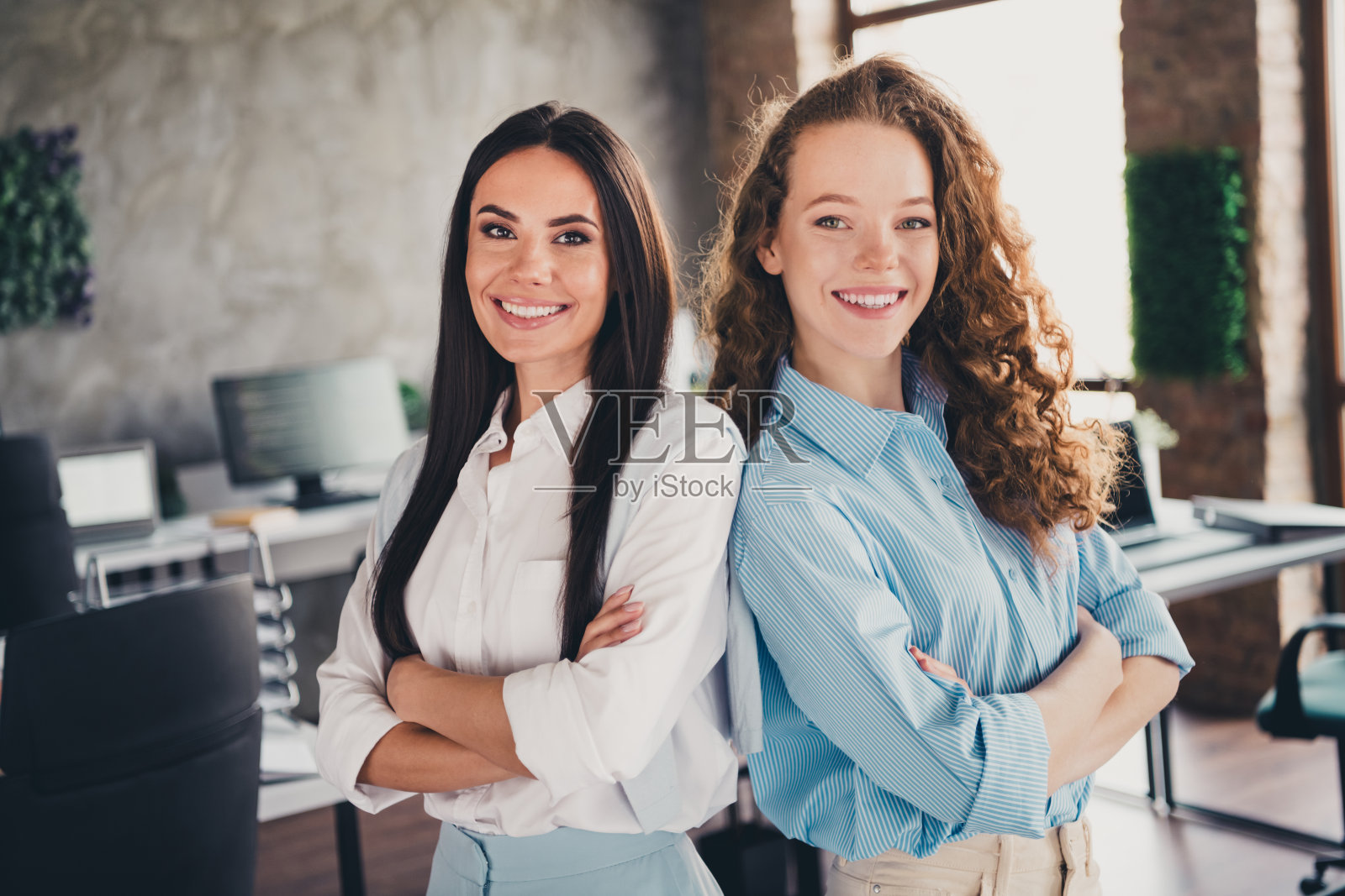 两位成功的迷人女性办公室工作人员自信地交叉双臂，站在室内工作场所。照片摄影图片