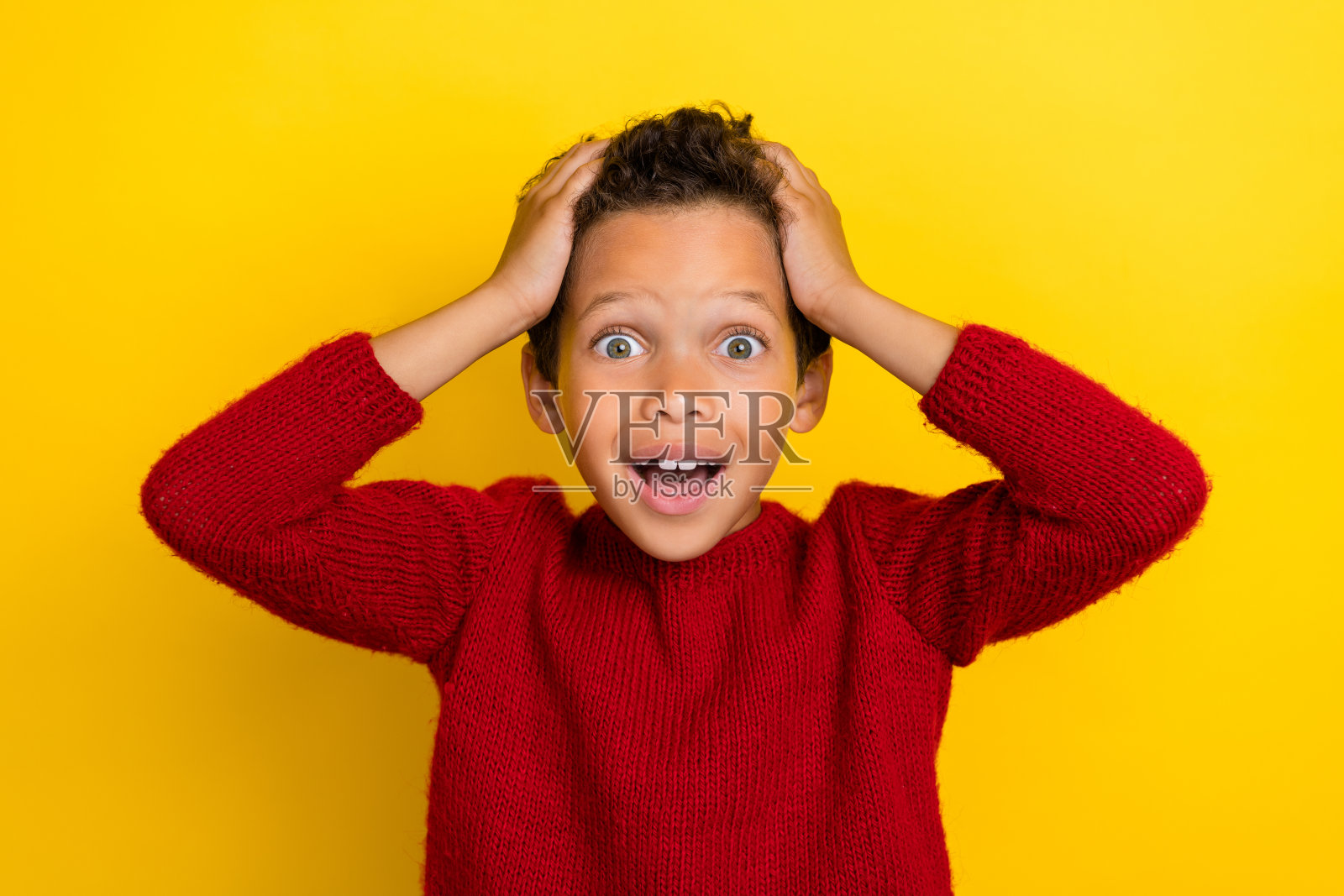 可爱的男孩触摸头部，难以置信自己的运气，穿着时尚的红色针织衫，背景为黄色。照片摄影图片