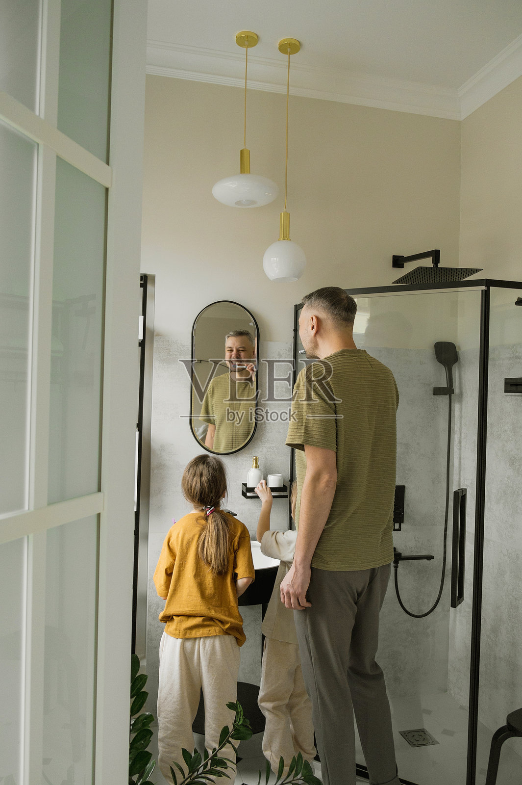 一位父亲在现代浴室里向他的两个小女儿演示正确的刷牙技巧。女孩们正在模仿他的动作，而父亲则在镜子里看着自己。照片摄影图片