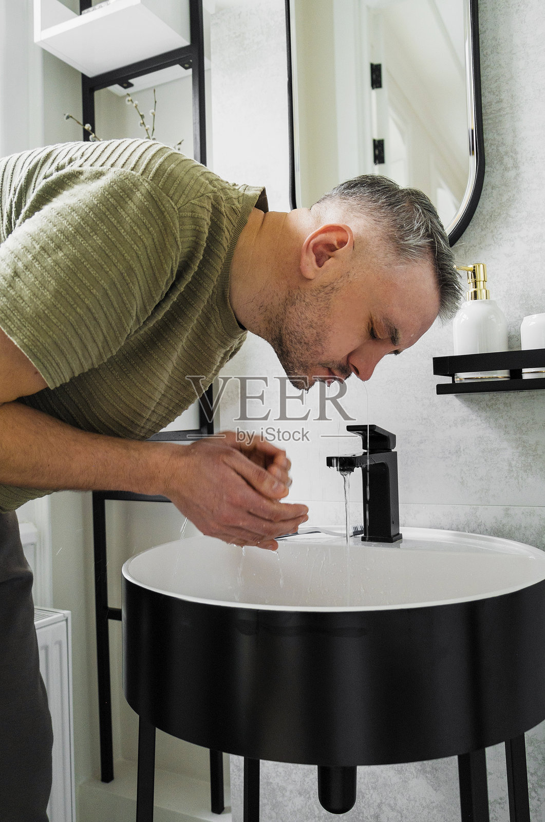 一名男子在现代浴室的洗手池前洗脸。他弯着身子，水从水龙头流出。照片摄影图片