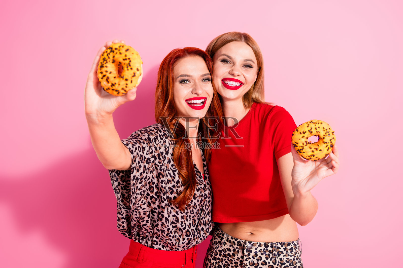 穿着红色豹纹时尚服装的美丽快乐女孩们拥抱在一起，手里拿着甜甜圈，背景是粉色的。照片摄影图片