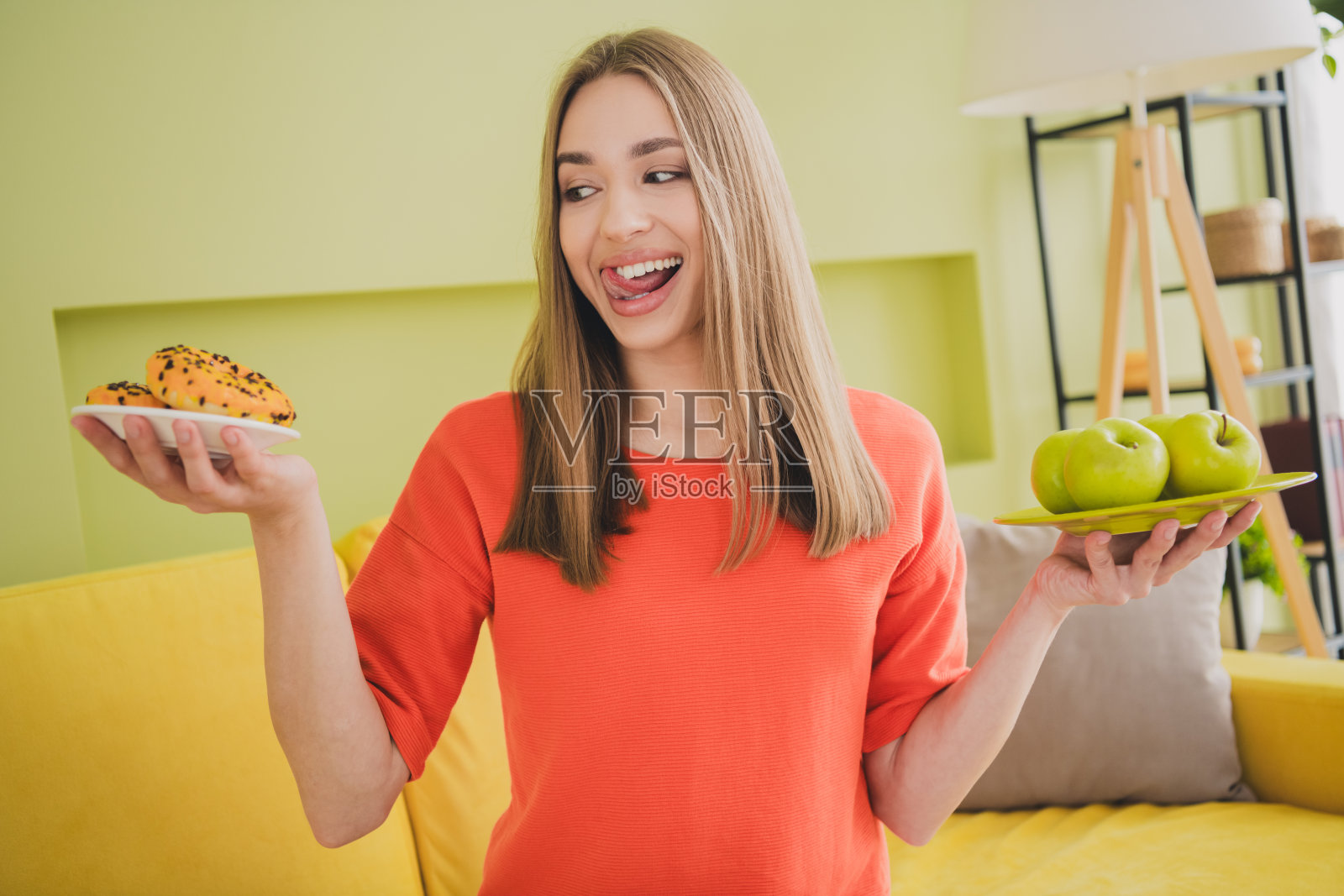 可爱的年轻女孩兴奋地比较苹果和甜甜圈，穿着橙色衣服，坐在宽敞的春季室内房间里。照片摄影图片
