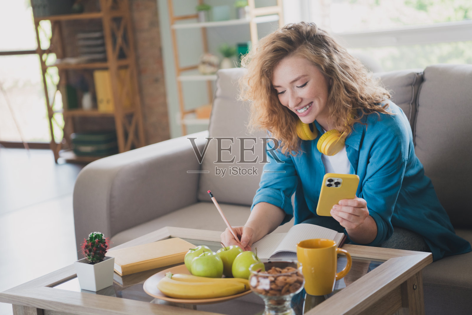 一位迷人的年轻红发女性穿着蓝色服装，坐在沙发上写笔记，享受温馨的日光家庭室内环境。照片摄影图片