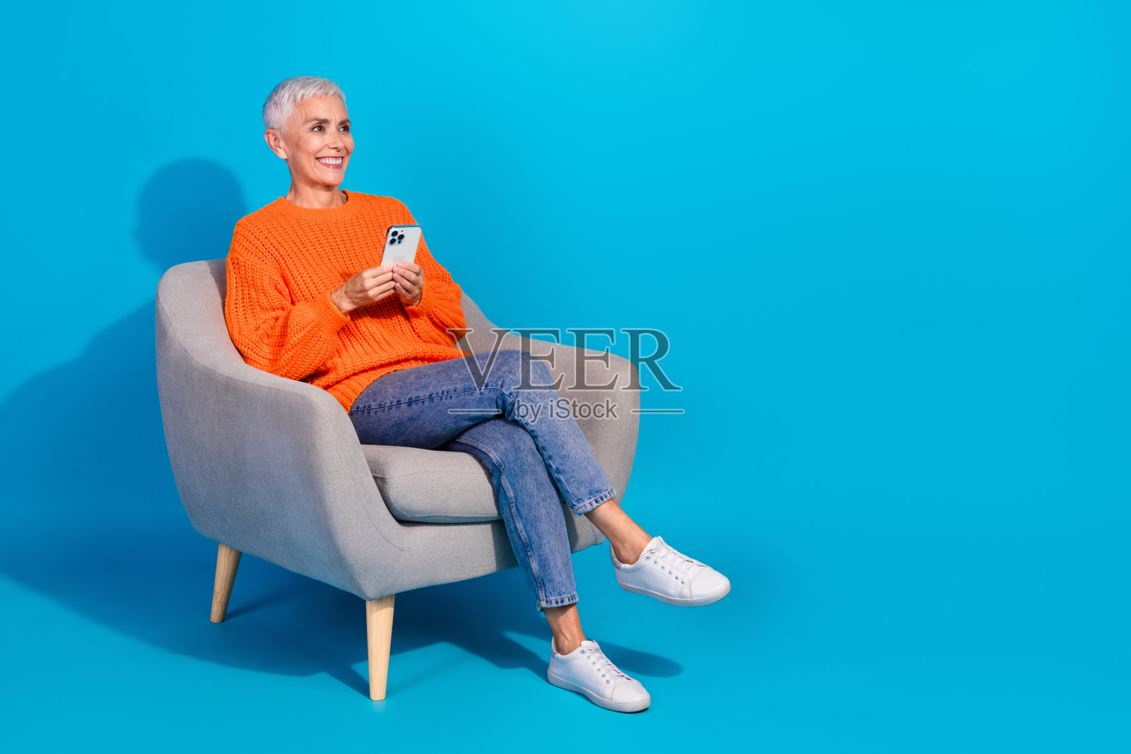积极的现代老奶奶坐在扶手椅上，使用电子设备，望向空旷的空间，背景为蓝色。照片摄影图片