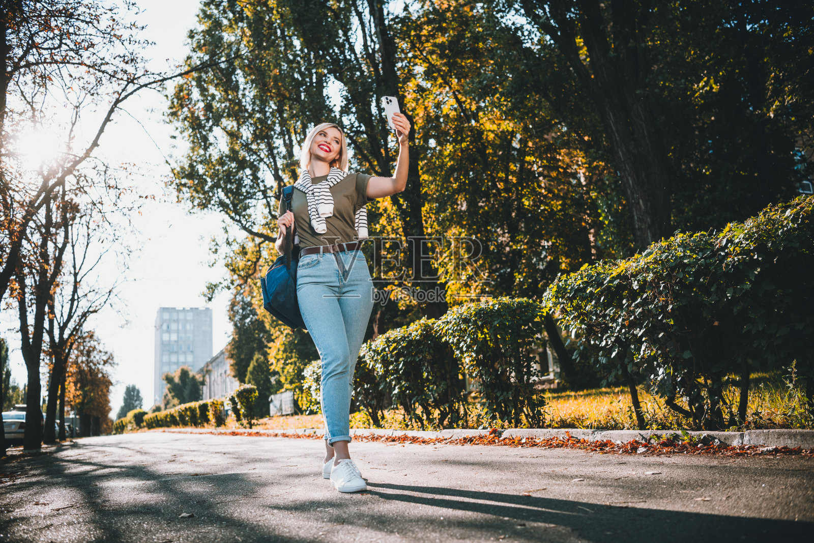 年轻漂亮的金发女子在城市公园外穿着卡其色衣服，肩上系着毛衣，拍全身自拍照。照片摄影图片
