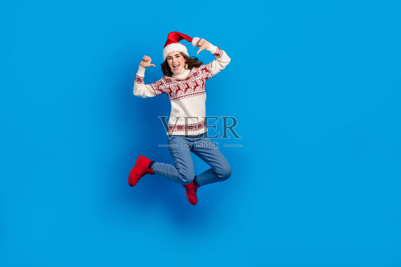 积极向上的酷女孩穿着毛衣，戴着圣诞帽，跳得很高，指着自己，背景为空旷的蓝色。照片摄影图片