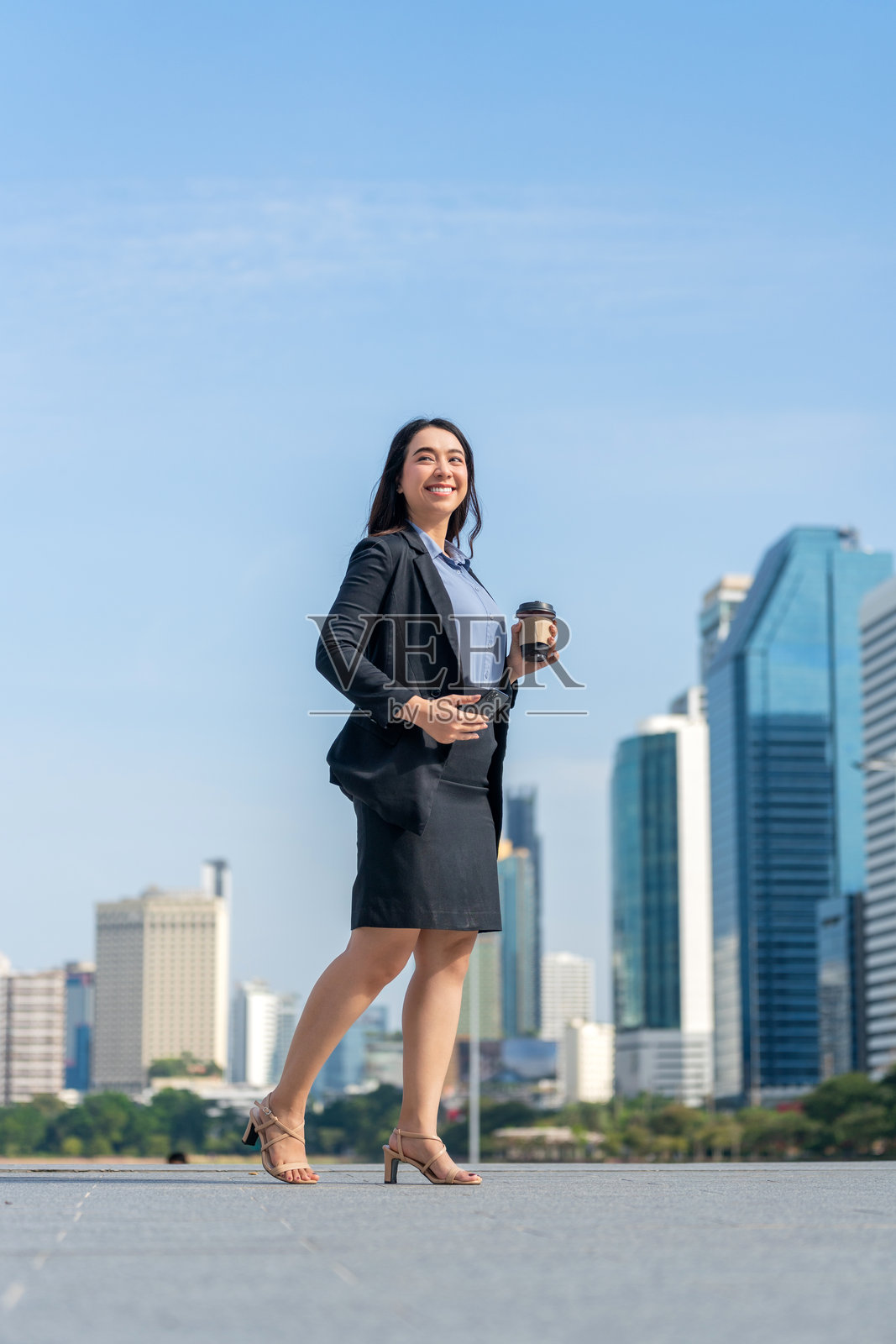 亚洲女商人使用智能手机，手持咖啡杯，背景是城市天际线照片摄影图片