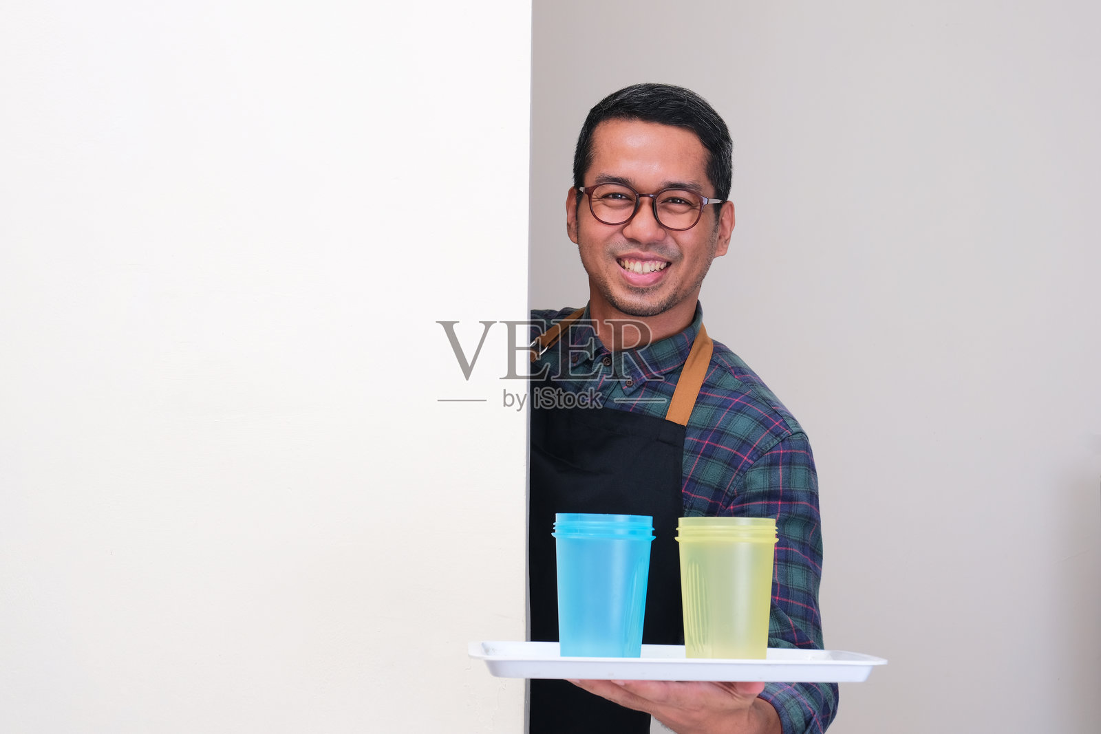 一个穿着围裙、面带笑容的男人从墙后探出头，手里托着两个饮料瓶。照片摄影图片