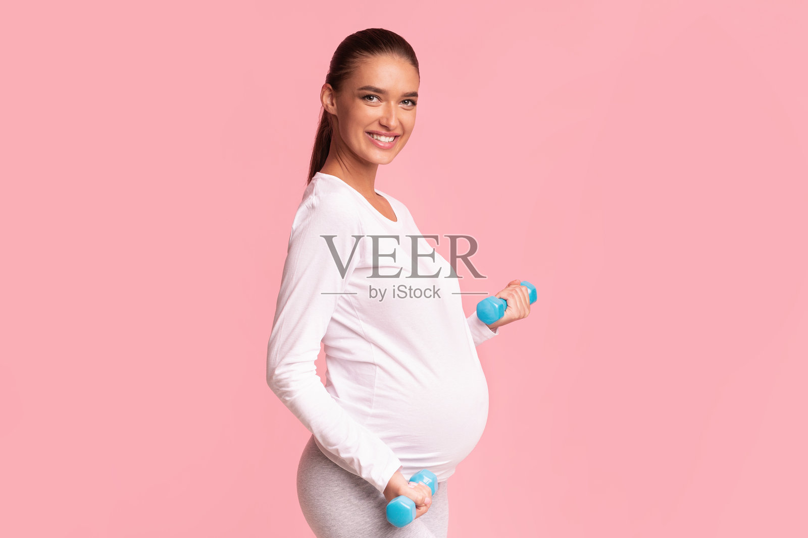 快乐的孕妇在粉色背景的工作室里举着哑铃照片摄影图片