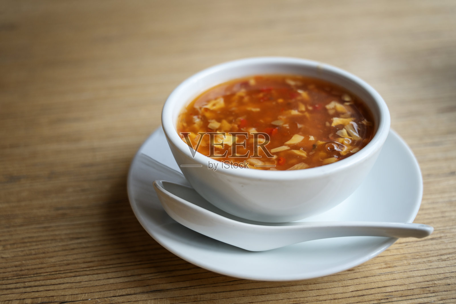 中式酸辣汤，汤中有轻盈的打散鸡蛋、肉类和蔬菜，盛在白色碗中，旁边有一只瓷勺，放在亚洲餐厅的木桌上，留有空白空间。照片摄影图片