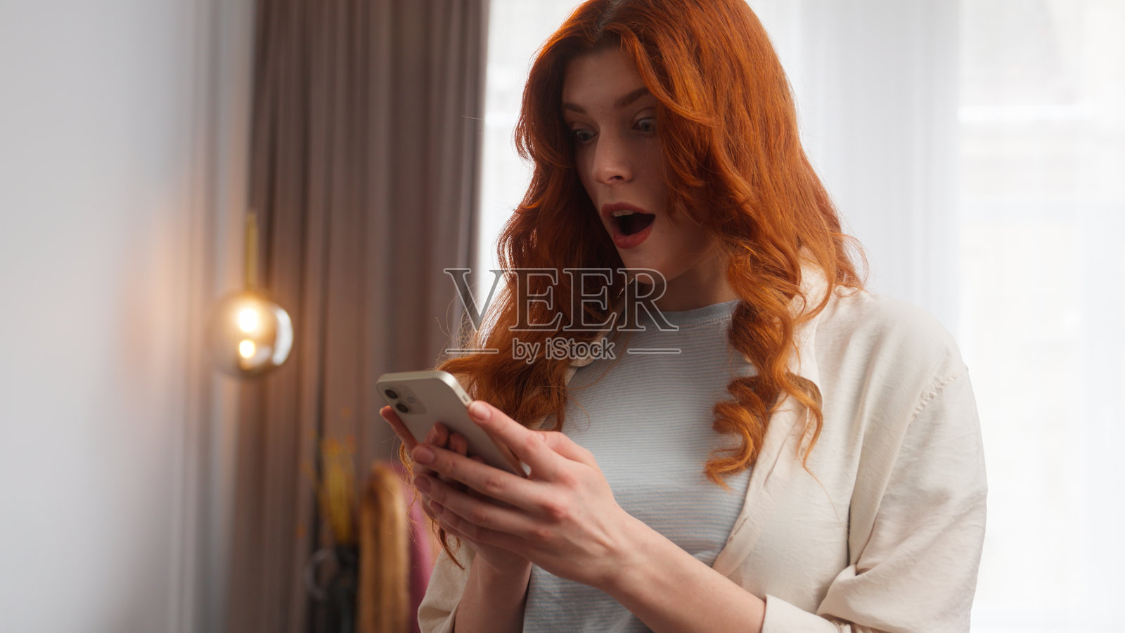 满意的白人女性惊讶地看着手机屏幕，开心地说“哇”，表现出惊喜、快乐和兴奋的情绪，红发女性微笑着，庆祝成功的好消息和优秀的结果，身处家中。照片摄影图片