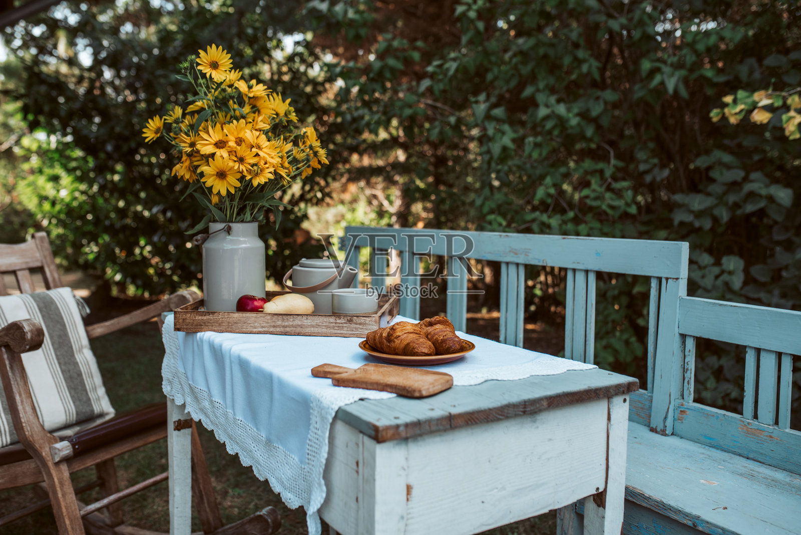 花园里树下的乡村风格桌子，上面铺着桌布，摆放着黄色花朵和食物的托盘。照片摄影图片