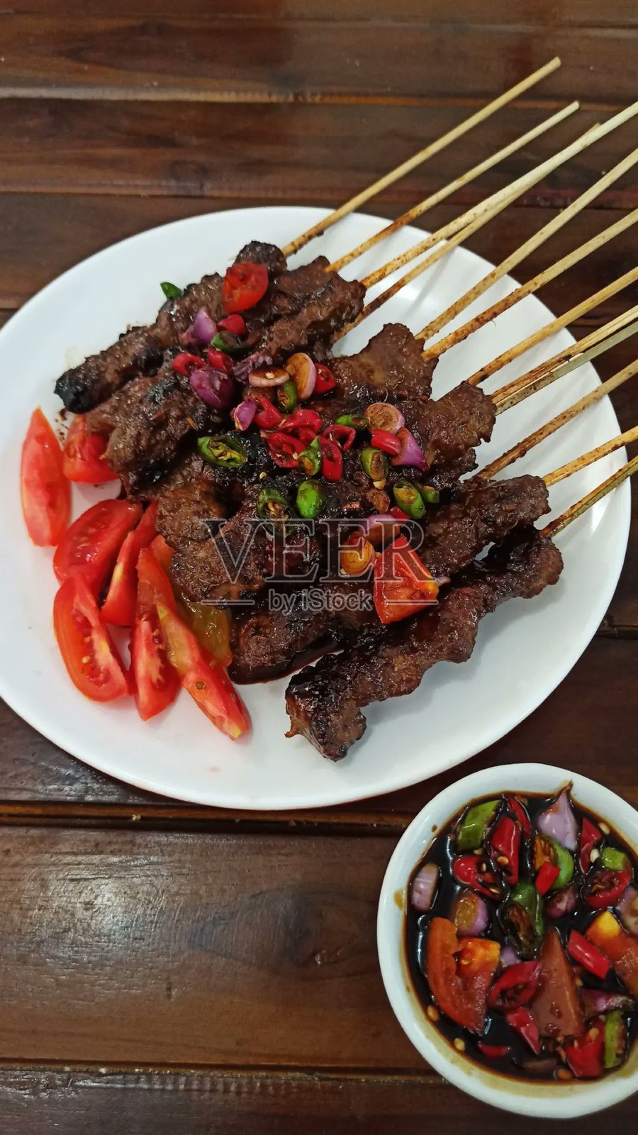 特写的羊肉沙爹（Sate Kambing），配有番茄、青葱、青辣椒和甜酱油，摆放在圆形白盘上。印尼传统美食。照片摄影图片