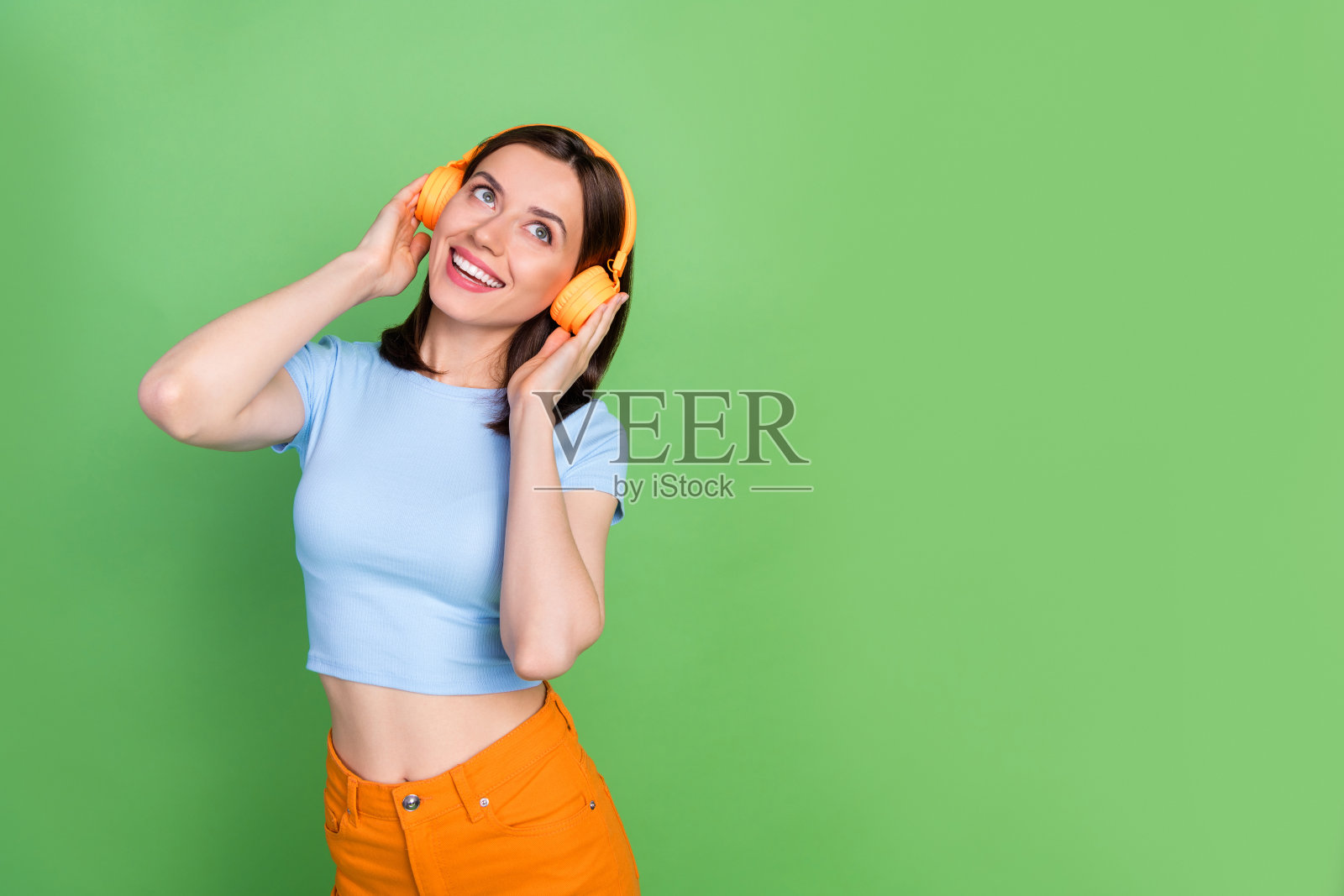 快乐迷人的女孩肖像，双臂触碰耳机，露出灿烂的笑容，目光投向空旷的空间，背景为绿色。照片摄影图片