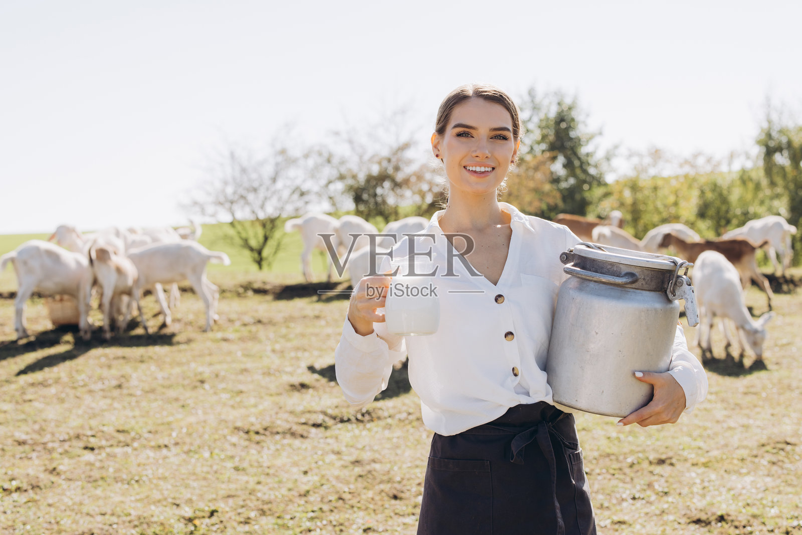 微笑的女性在阳光明媚的乡村山羊农场里手持新鲜牛奶壶和罐子照片摄影图片