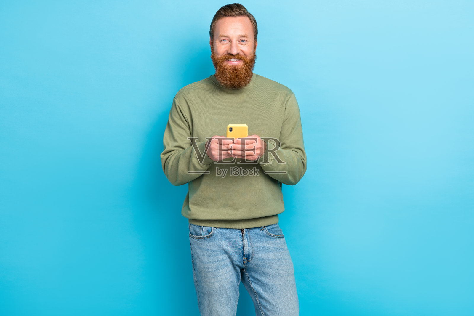 积极乐观的男士，红色发型，穿着卡其色长袖，手持智能手机，背景为青色。照片摄影图片