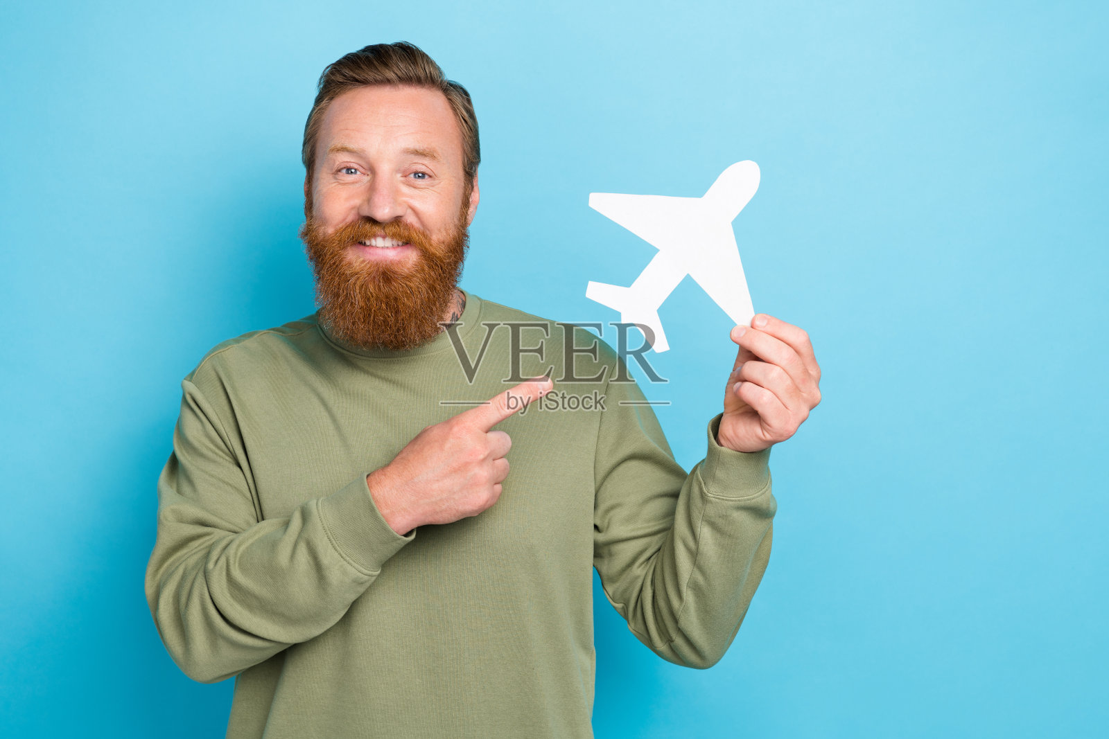 一位满意的金发男士穿着卡其色长袖衬衫，指着一架白色纸飞机，背景为蓝色。照片摄影图片