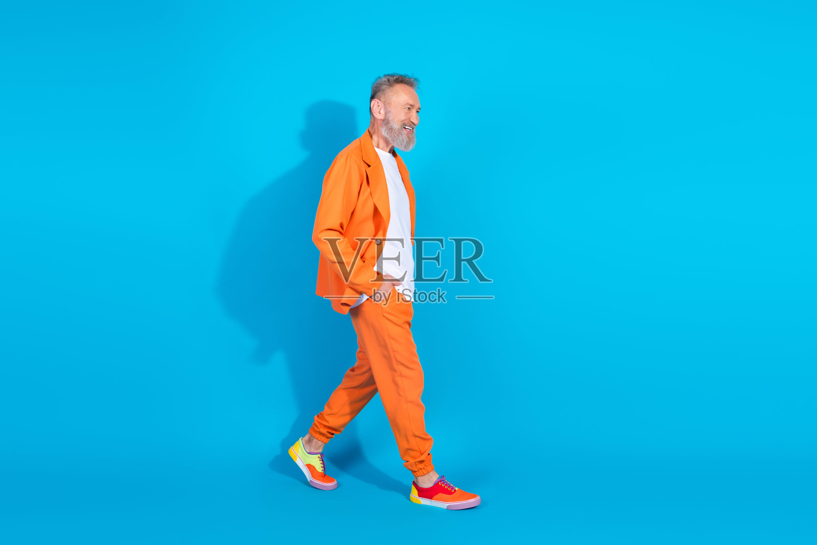 现代时尚老年男子全身侧面肖像，穿着橙色服装在蓝色背景下走动，周围空旷。照片摄影图片