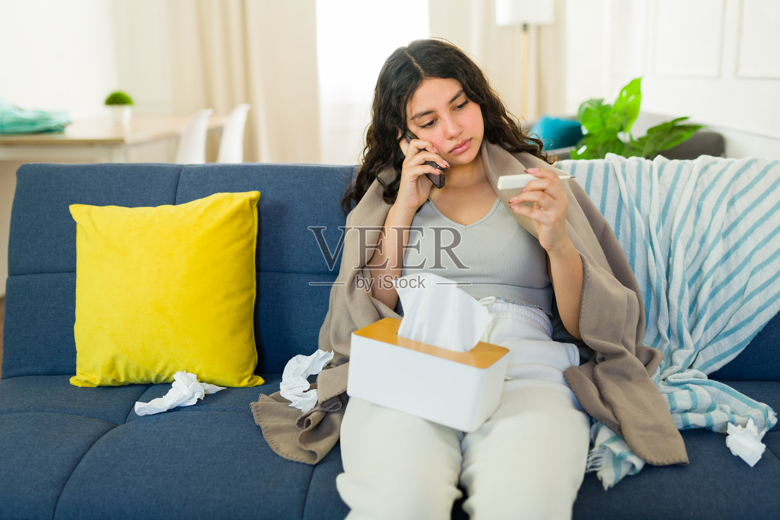 生病的青少年裹着毯子，坐在家里的沙发上用手机查看体温。照片摄影图片