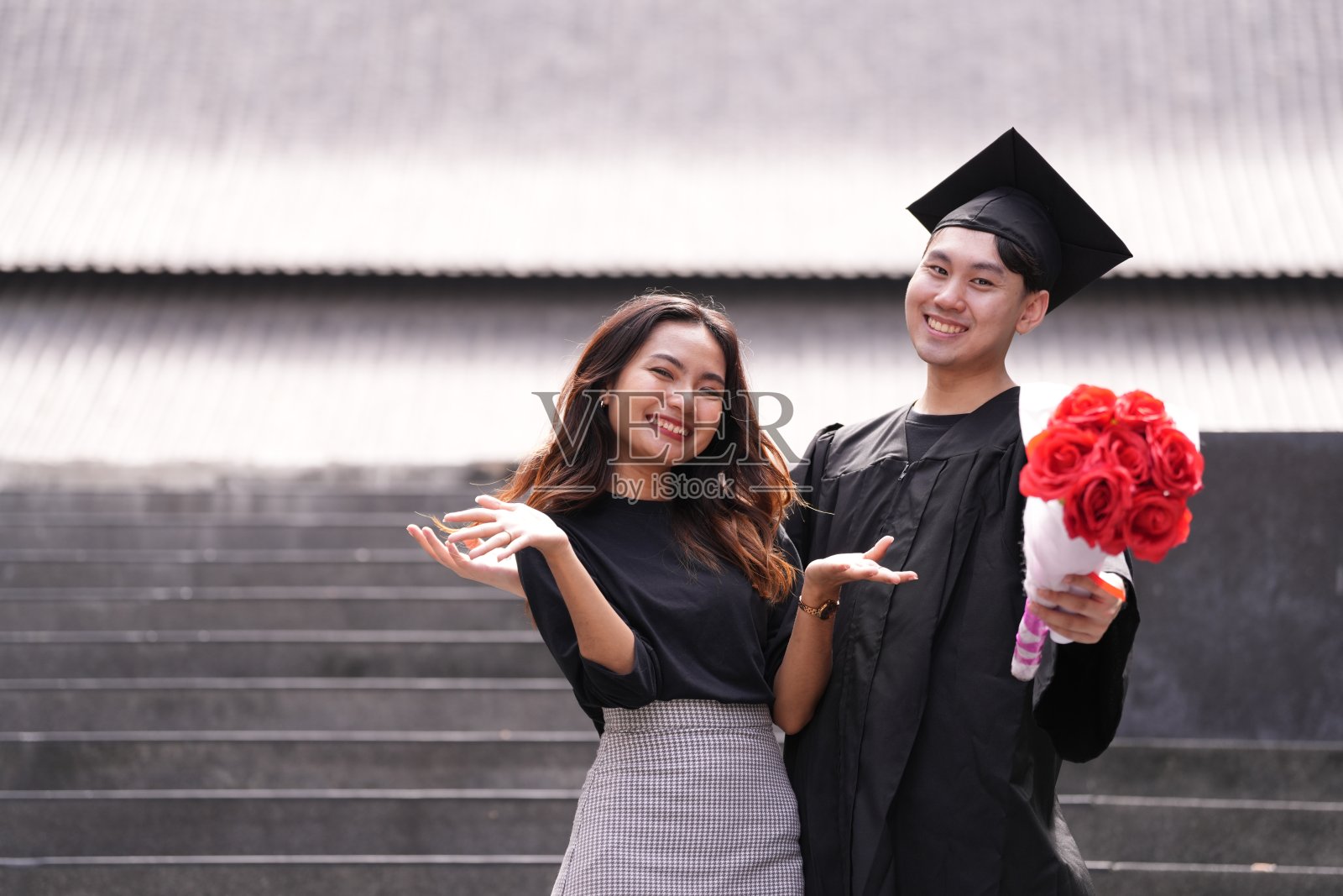 一位自豪的毕业生身穿学位服，手捧一束红玫瑰，站在微笑的朋友旁边。他们在现代背景的台阶上愉快地摆姿势，共同庆祝这一难忘的成就。照片摄影图片