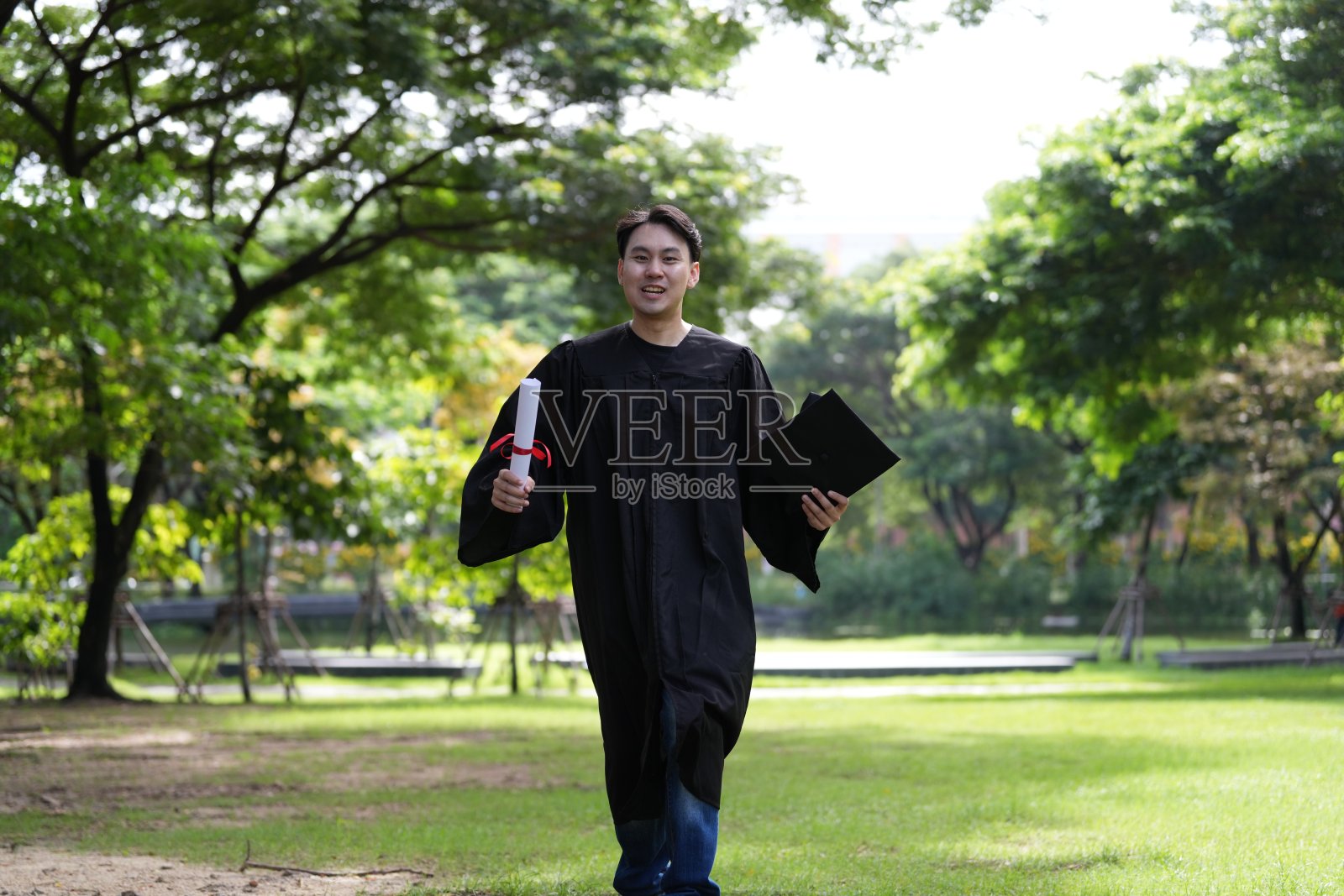 一位身穿黑色毕业袍的快乐毕业生在绿色的田野上奔跑，手里拿着毕业证书和文件夹，庆祝他的成就。背景是郁郁葱葱的公园环境，展现了兴奋和自豪。照片摄影图片