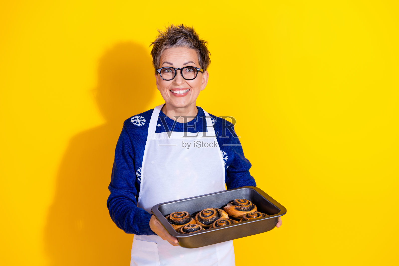 可爱的迷人老年女士穿着蓝色时尚服装，手持新鲜烘焙食品，背景为黄色。照片摄影图片